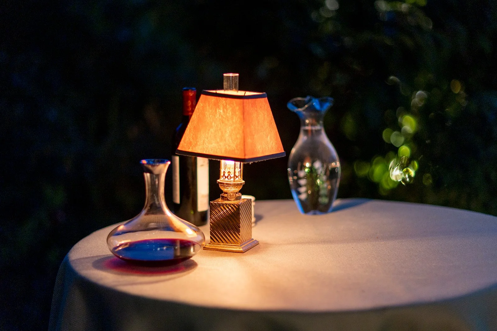 A table with a small orange lampshade, two glass decanters, and a wine bottle, set outdoors at night.