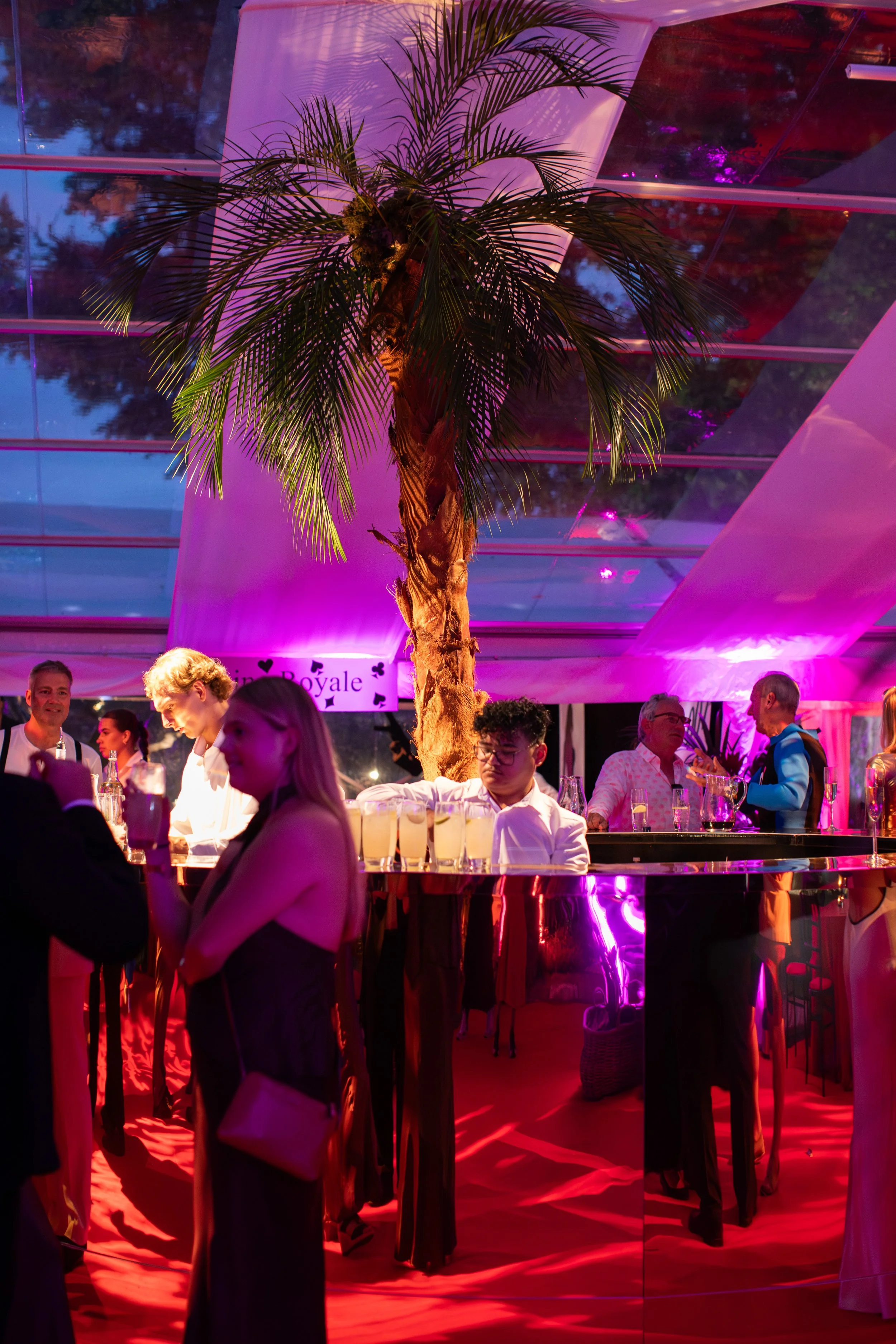 A lively indoor event with a tall palm tree under pink and purple lighting, guests dressed in formal attire socializing, drinks on the bar, and a decorative sign reading 'RoyaLe' in the background.
