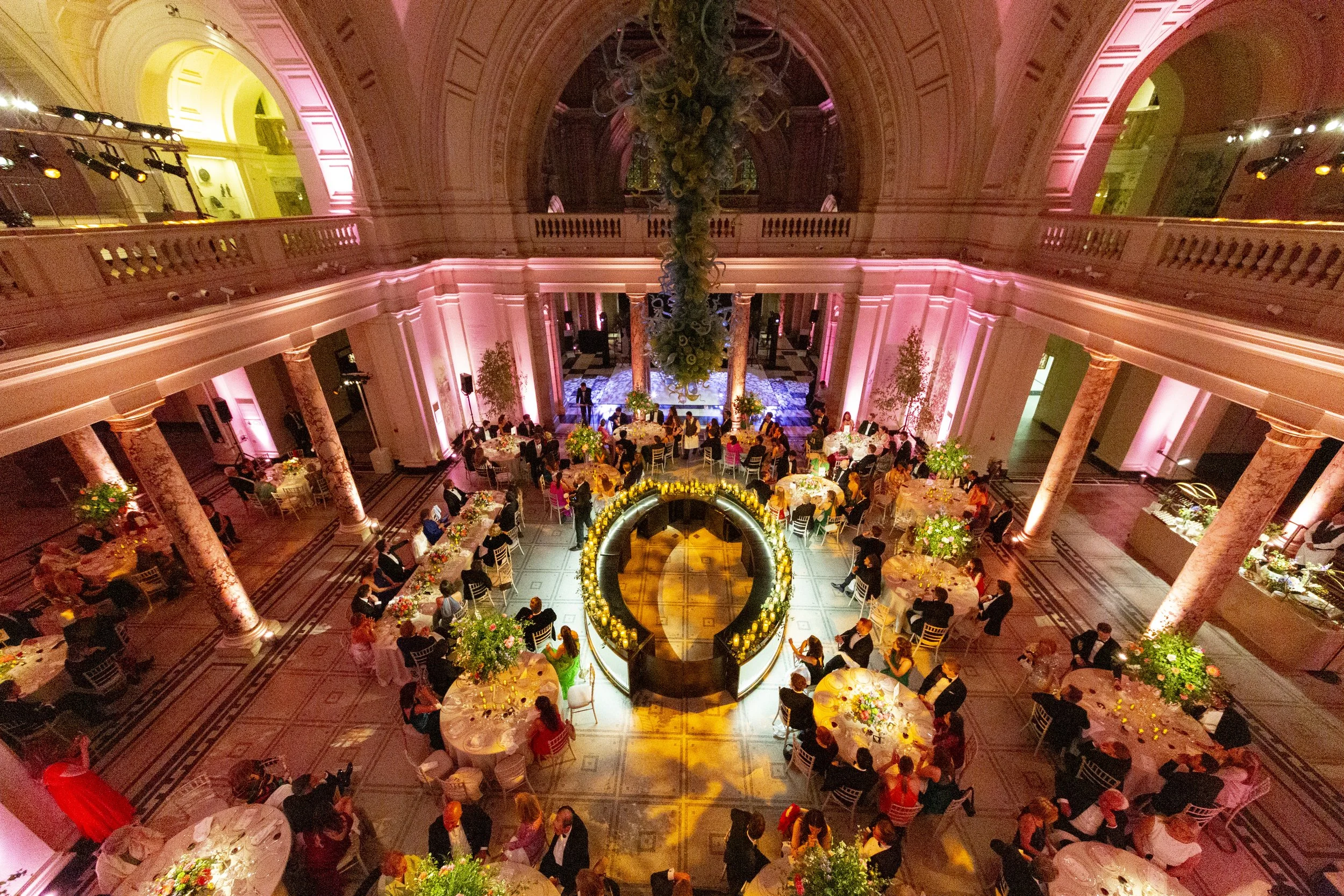 Elegant ballroom decorated with pink lighting, featuring a chandelier, a floral centerpiece, and round tables with guests dining. Columns and two-tiered balcony overlook the spacious area.