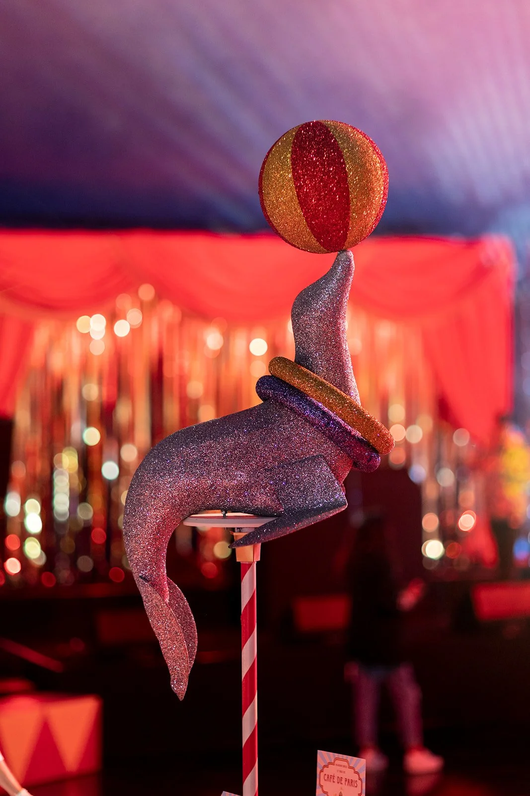 A glittery pink flamingo sculpture balancing a colorful striped bouncy ball on its beak, with a blurred red and pink background of circus or carnival tent decorations.