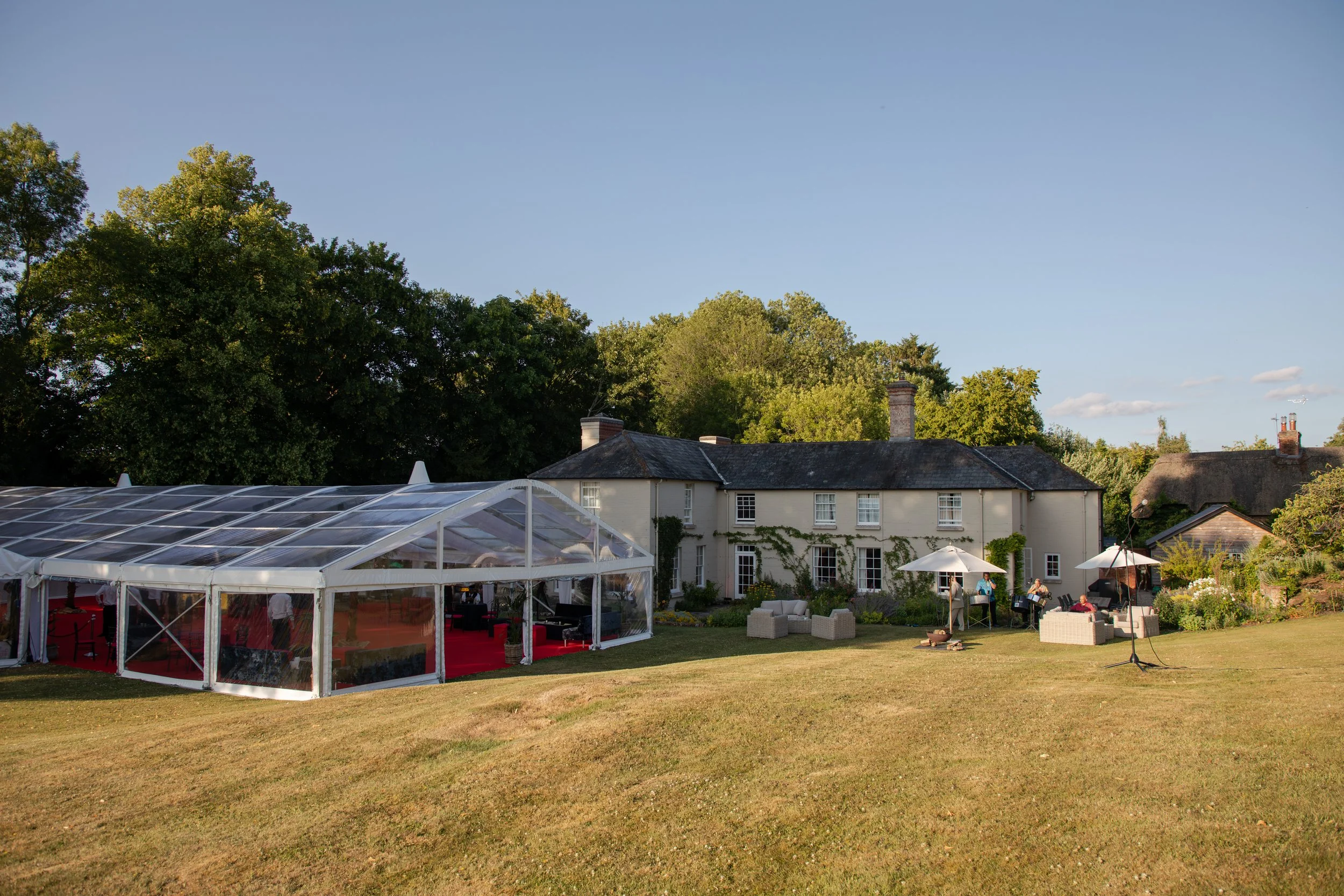 An outdoor garden party scene with a white house, green trees, and a large glass tent on the grass, with people gathered under umbrellas and seating areas.