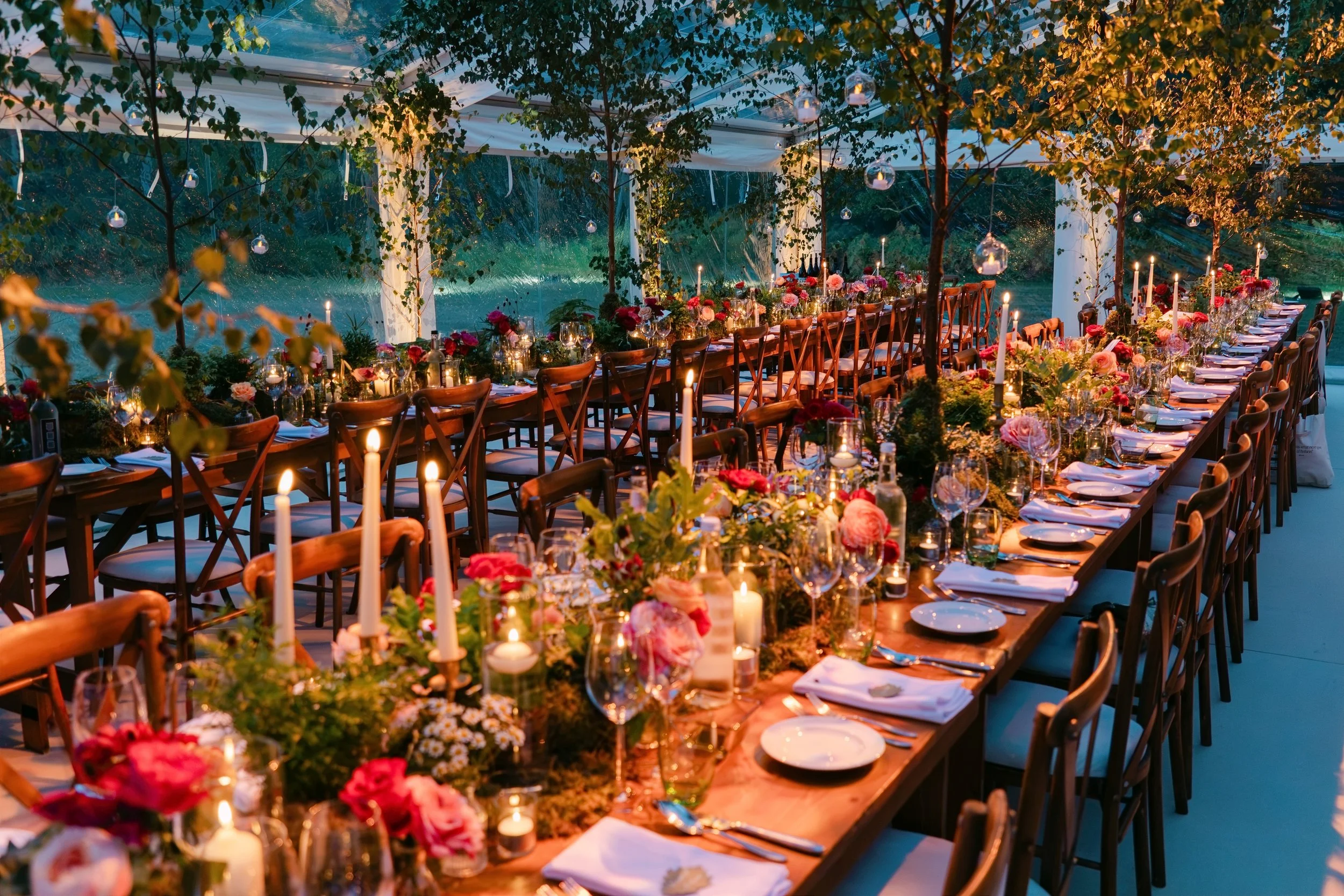 A long dinner table set for an outdoor celebration, decorated with flowers, candles, and glassware, inside a transparent tent with string lights and surrounded by trees.