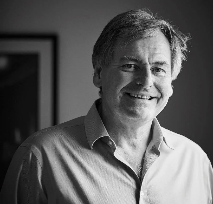 A smiling middle-aged man with light hair wearing a collared shirt standing indoors.