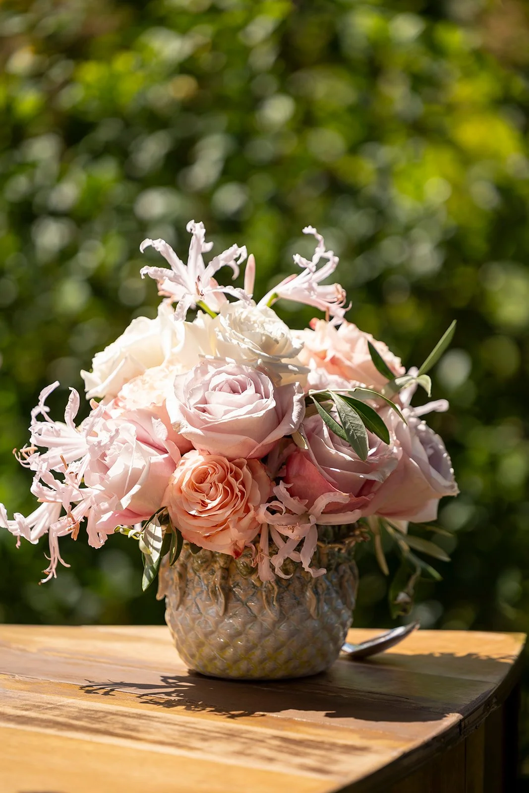 A bouquet of pink and white roses and lilies in a decorative ceramic vase placed on a wooden surface outdoors with a blurred green background.