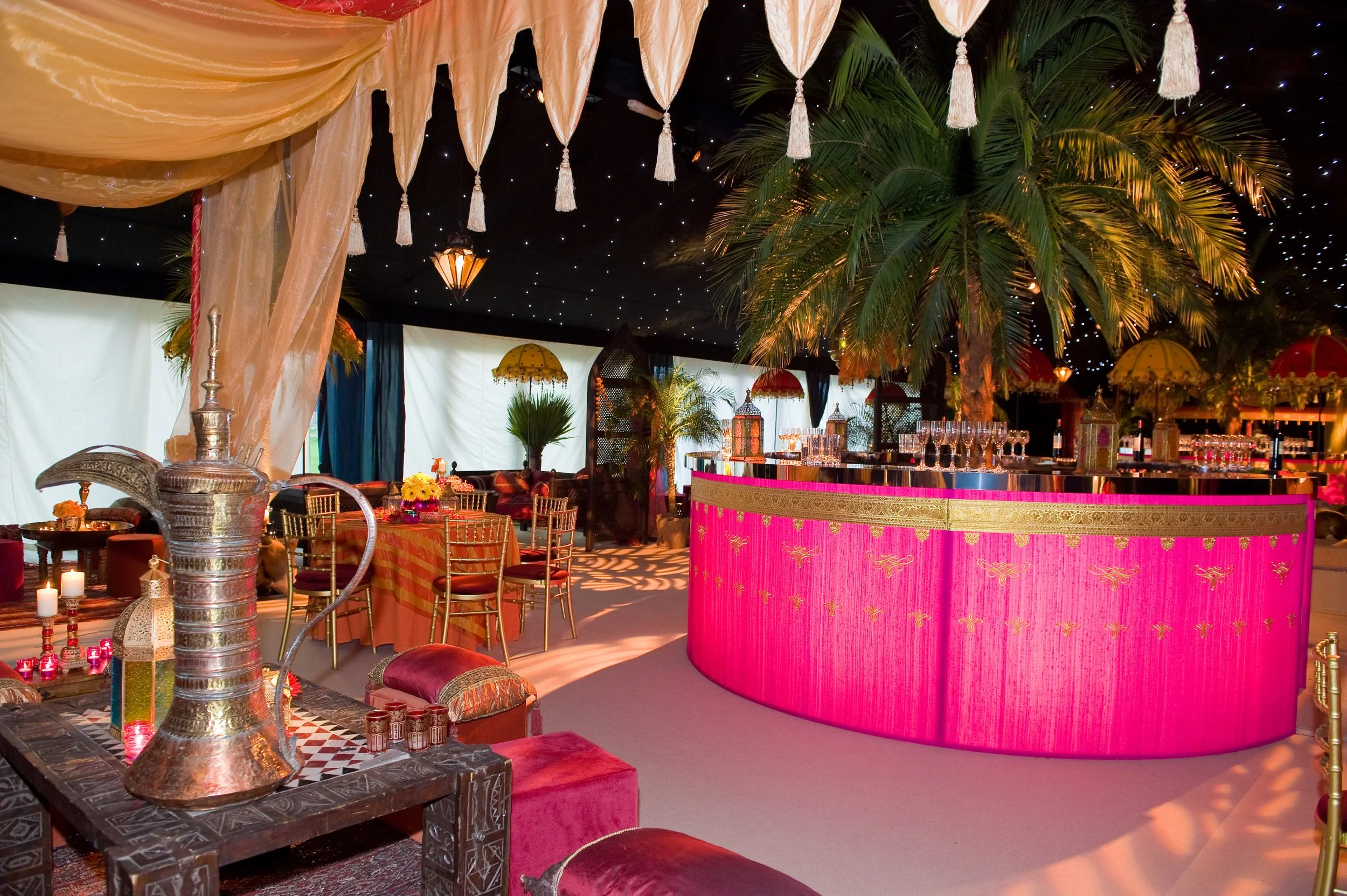 Interior of a decorated event space with seating, ornate drapes, a pink bar, lush green palm trees, decorative lanterns and candles, and a starry black ceiling.