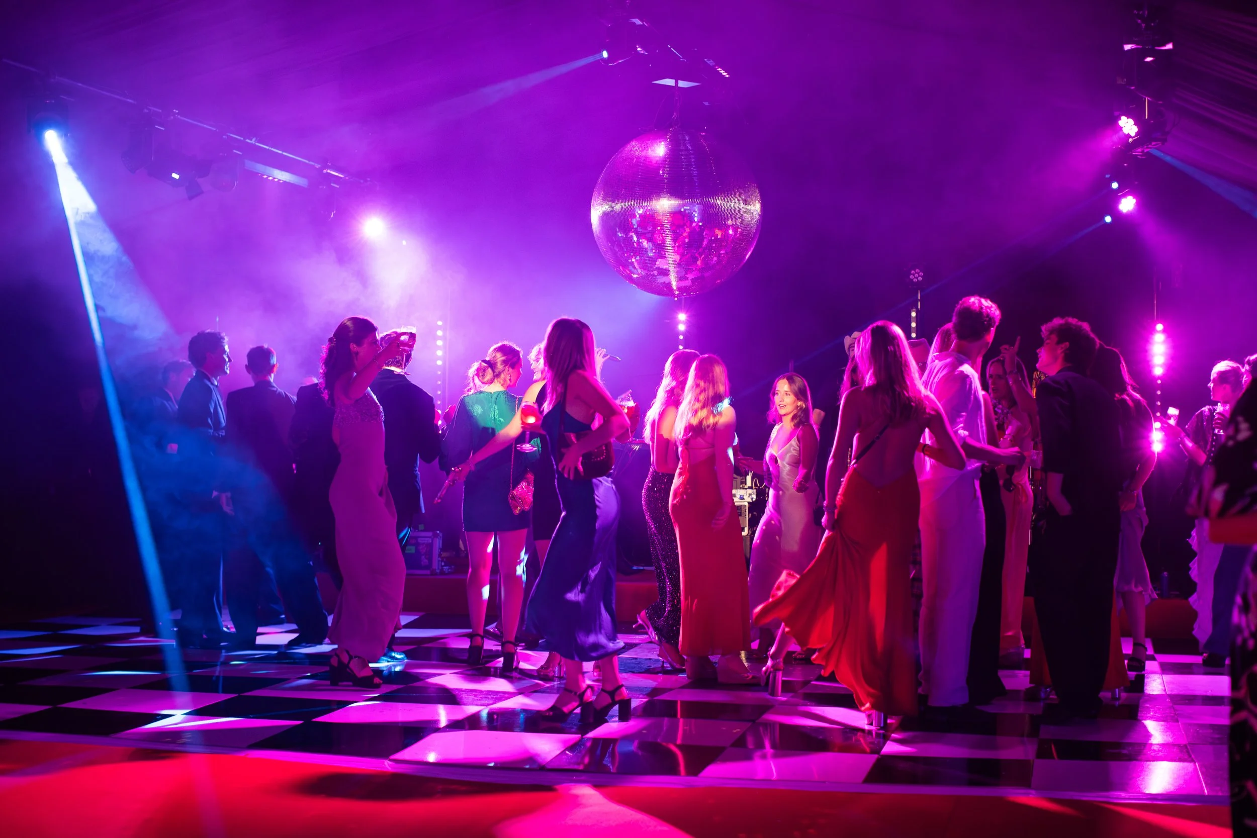 A group of people dancing on a checkered dance floor under purple and pink lighting with a large disco ball hanging from the ceiling.
