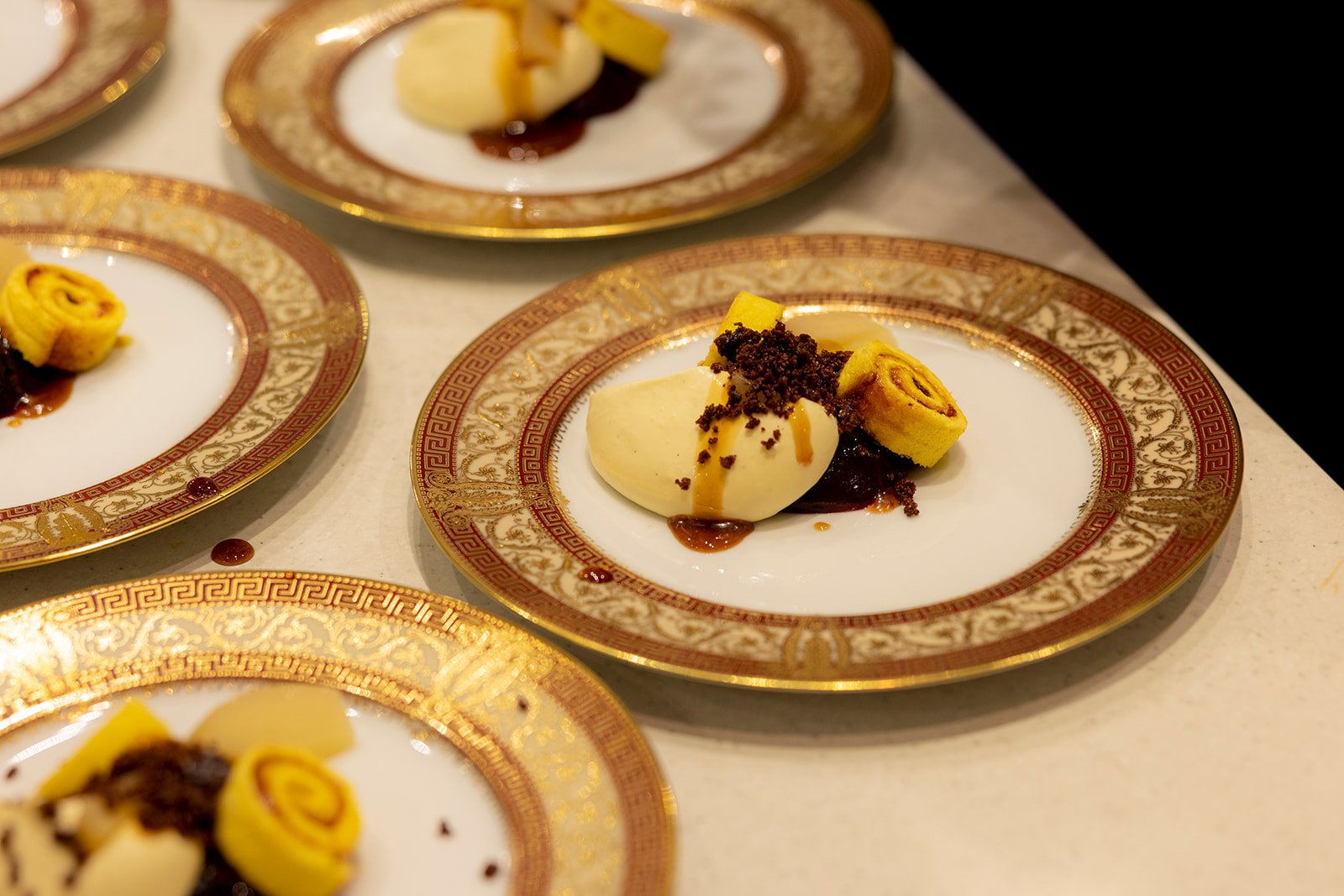 Elegant dessert plates with mousse, rolled cake, and chocolate crumbs on ornate gold and red patterned china.