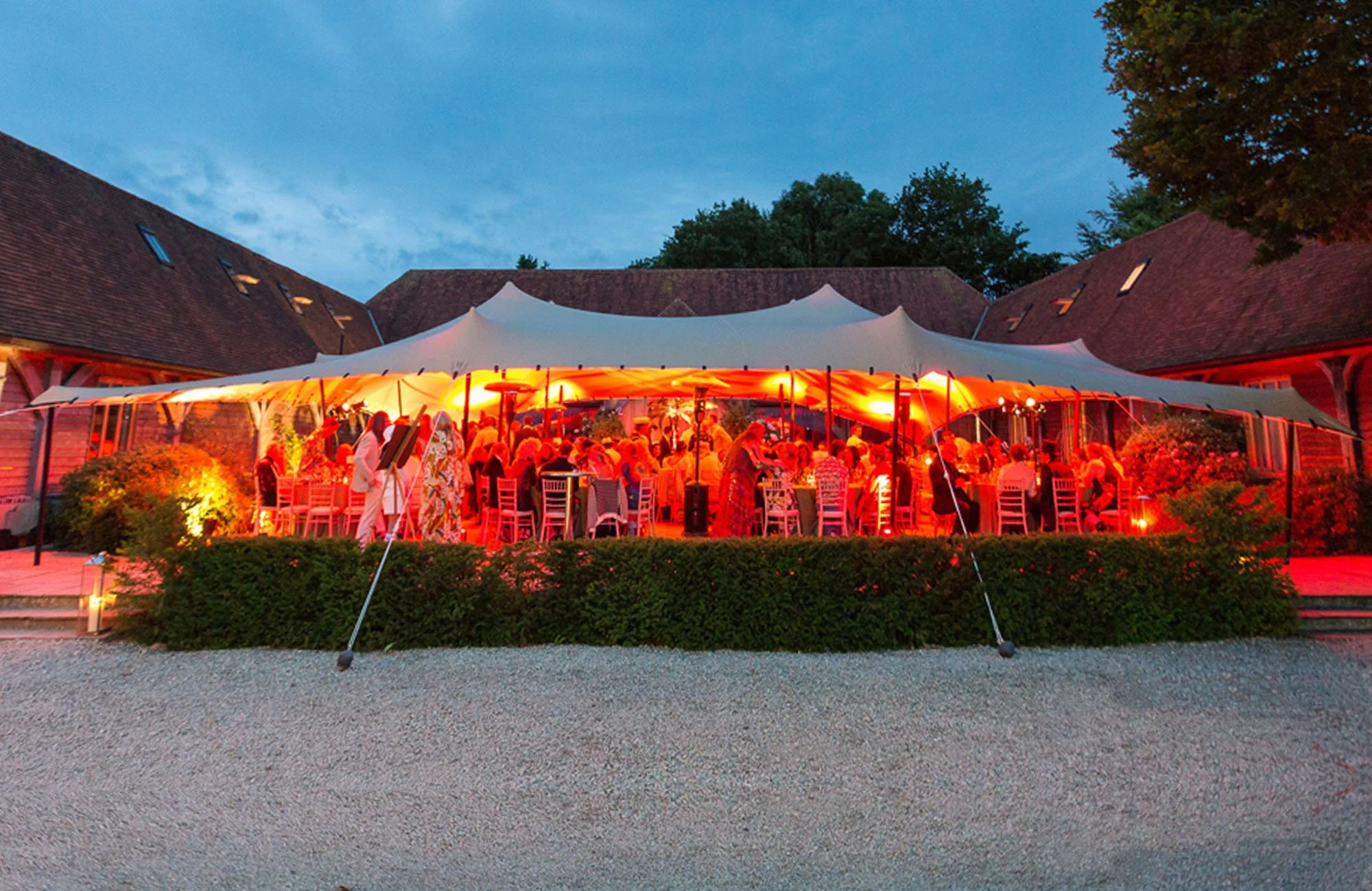 An outdoor event at dusk under a large white tent with warm lighting, surrounded by brick buildings and trees, guests seated at tables enjoying a social gathering.