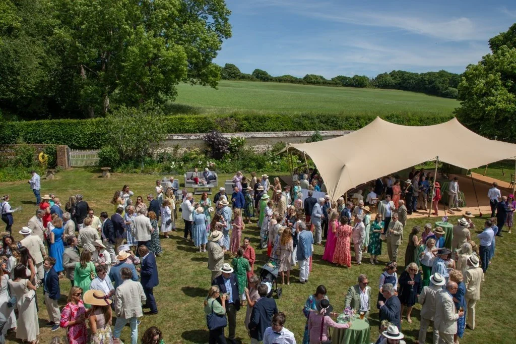 Outdoor wedding or celebration with many guests gathered on a grassy lawn under a large cream-colored canopy, with a scenic green landscape in the background.