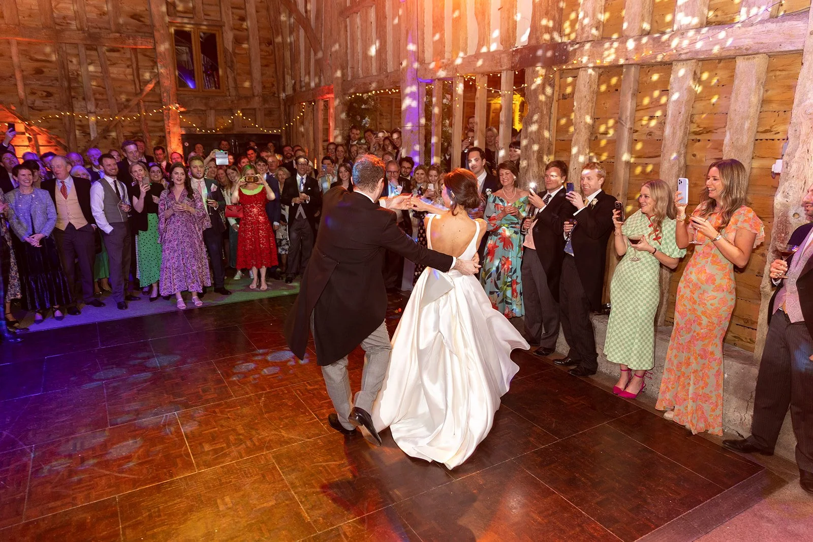Bride and groom dancing at their wedding reception, with guests watching and taking photos in a rustic wooden venue.