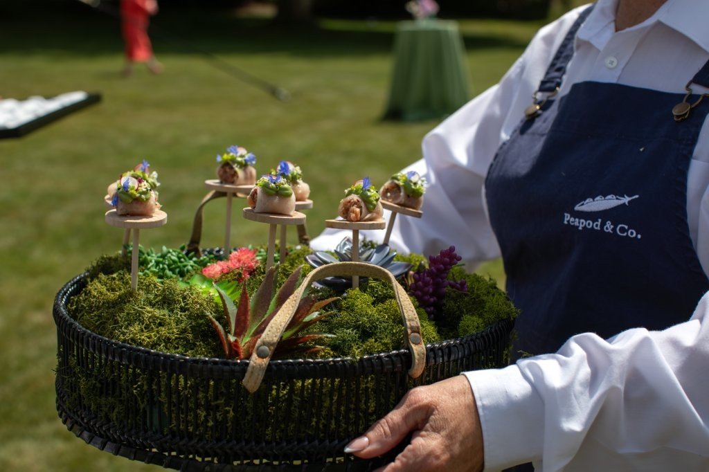 A person holding a black basket filled with moss and small decorative plants, with a few miniature food items or appetizers on small wooden platforms above the moss, decorated with edible flowers.