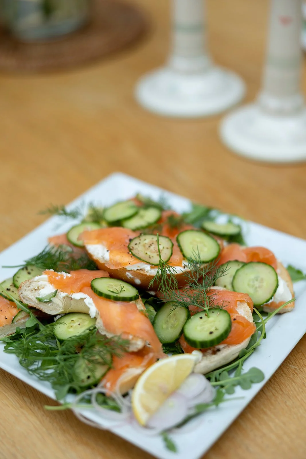 Open-faced smoked salmon bagel with cucumber slices, fresh herbs, lemon wedge, and cream cheese on a white plate.