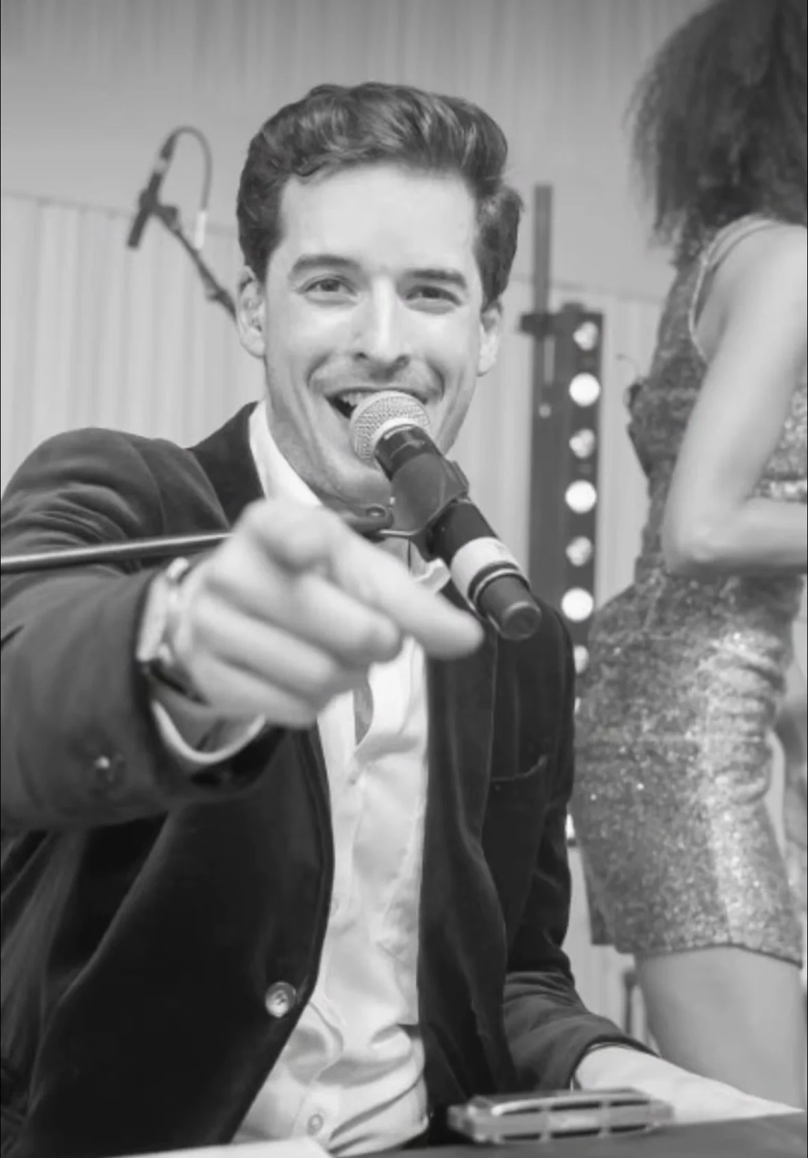 A young man with dark hair wearing a suit jacket and white shirt, smiling and pointing at the camera, sitting at a table with a microphone in front of him. A woman with curly hair, wearing a glittery dress, is standing beside him, partially visible. The photo appears to be taken at an event or celebration.