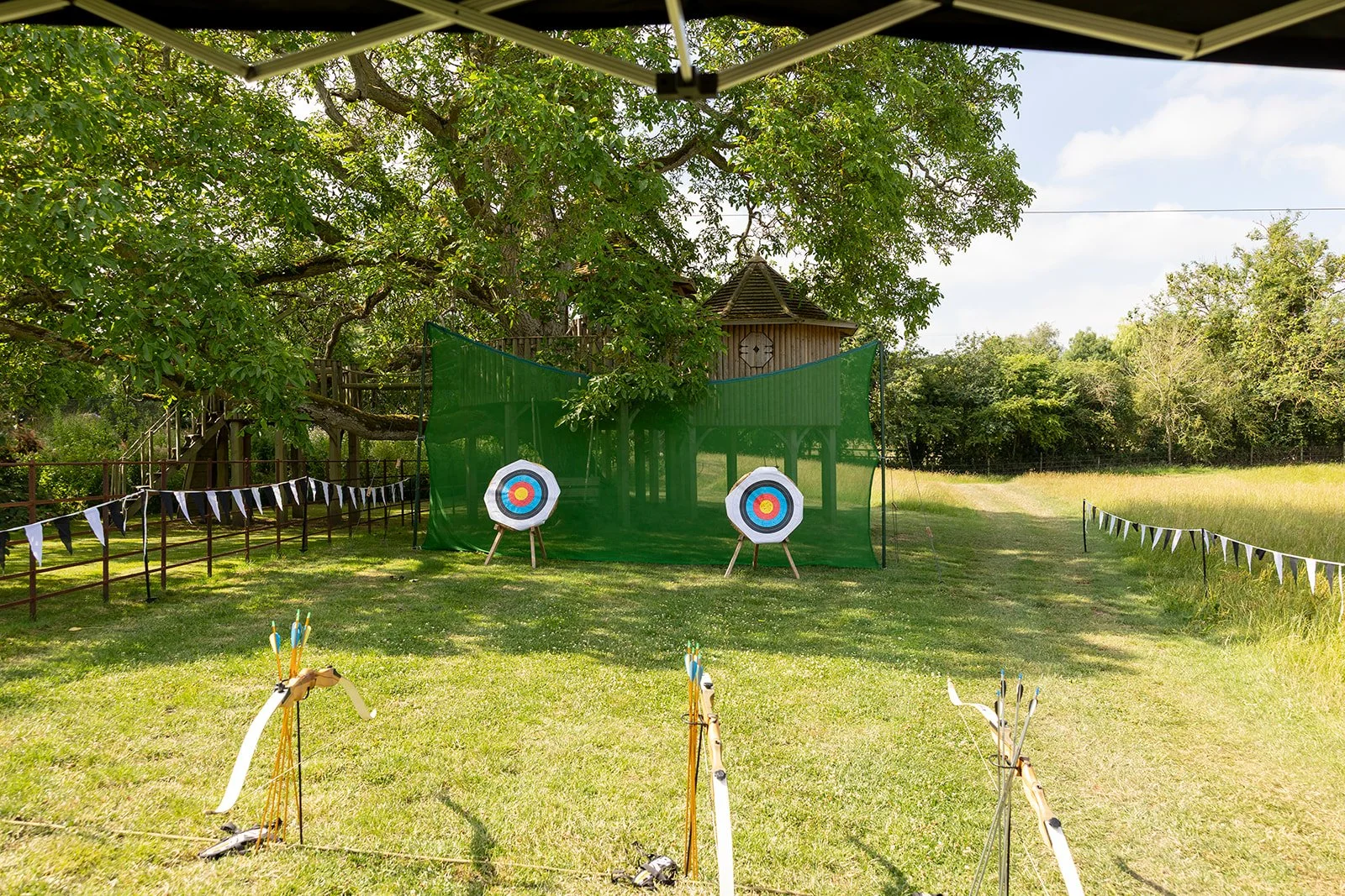 Archery targets set up outdoors on grass with bows and arrows in foreground, green netting and wooden treehouse in background, surrounded by trees and open field under blue sky.