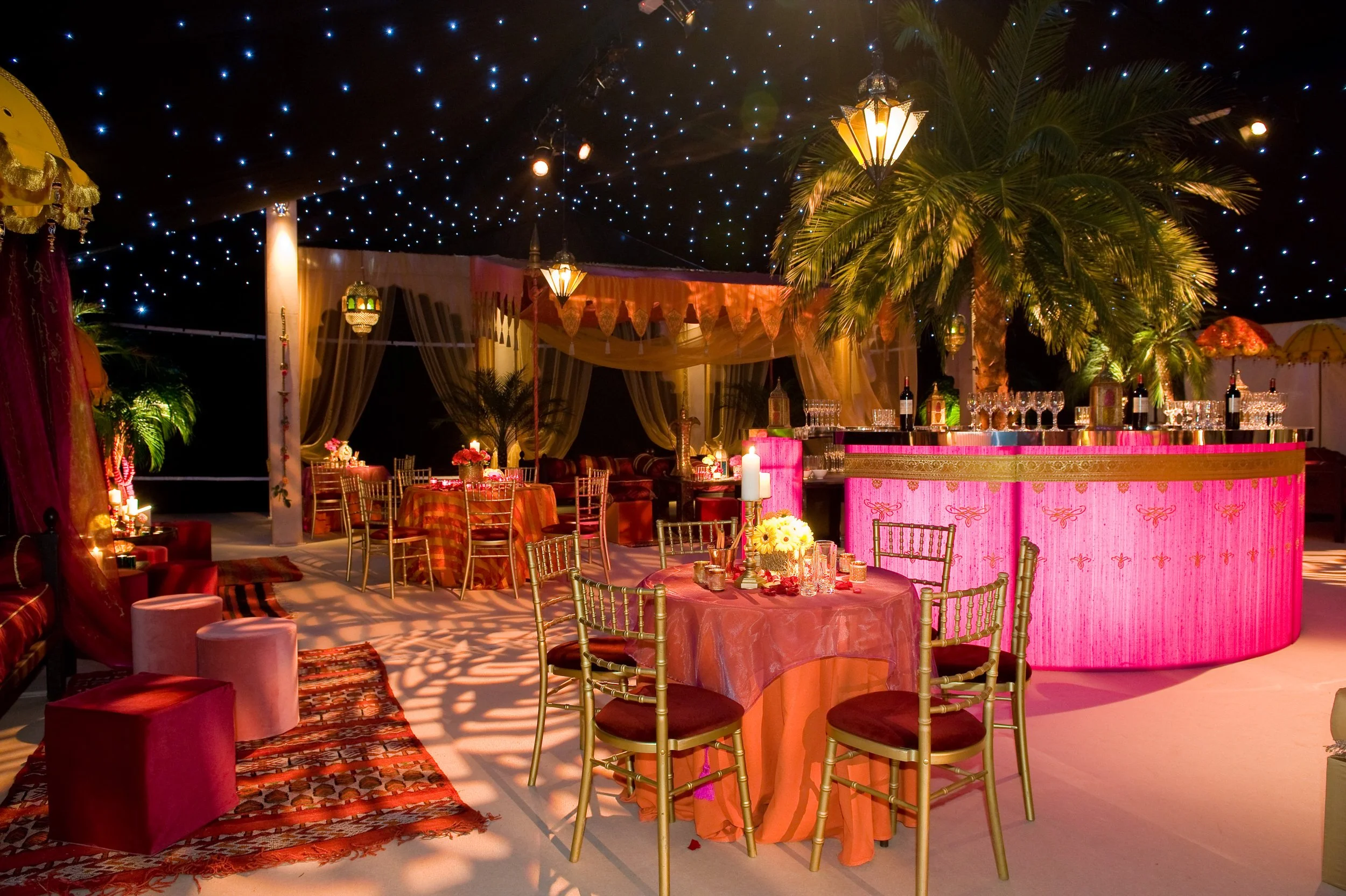Indoor banquet setup with round tables covered in pink tablecloths, decorated with flowers and candles, surrounded by gold chairs. A pink bar with glasses and bottles is in the background, with palm trees and ornate lanterns. The ceiling is decorated