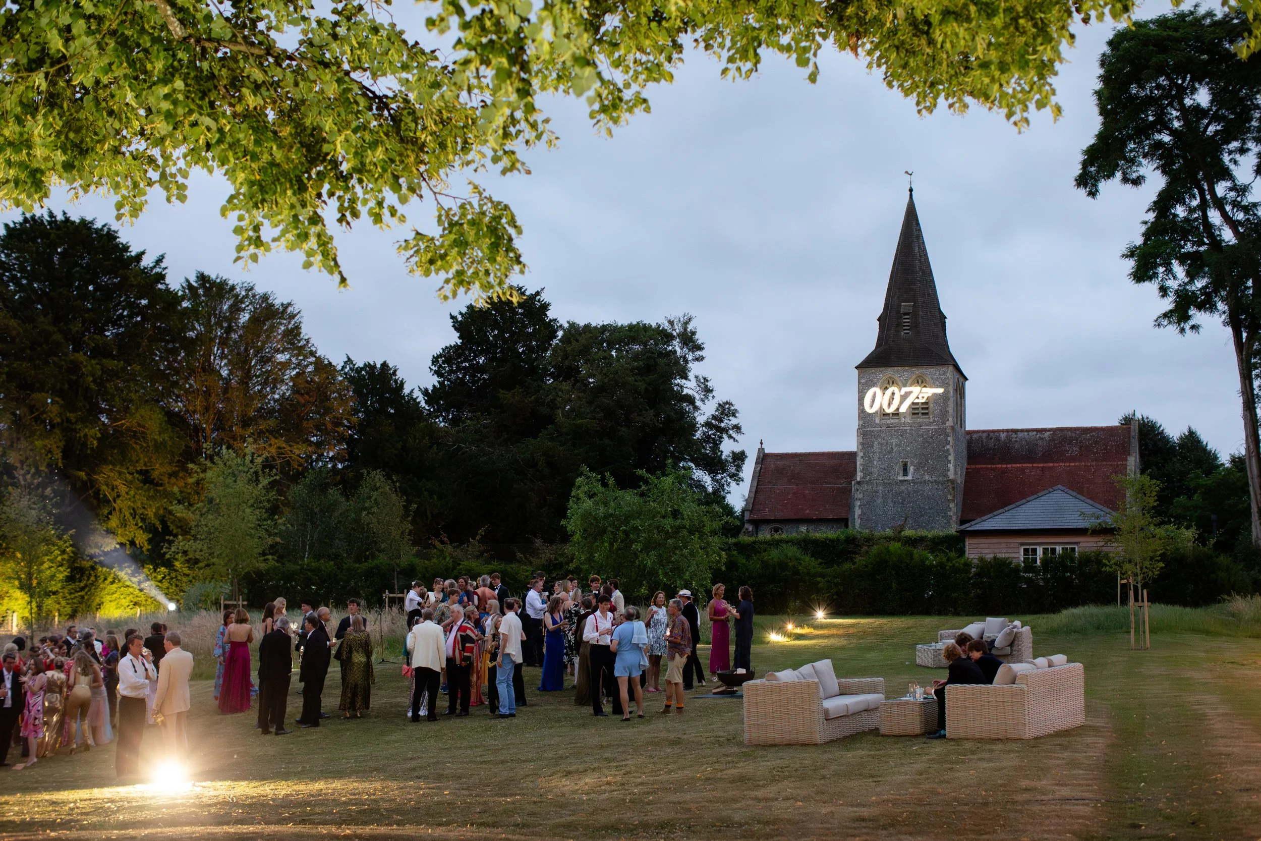 Outdoor wedding reception in a garden with guests, white chairs, and a historic church steeple in the background during sunset.