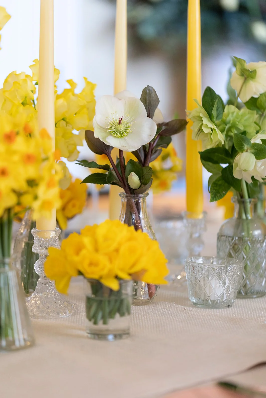 Various vases with yellow and white flowers, including daffodils and other blossoms, on a table with yellow candles.