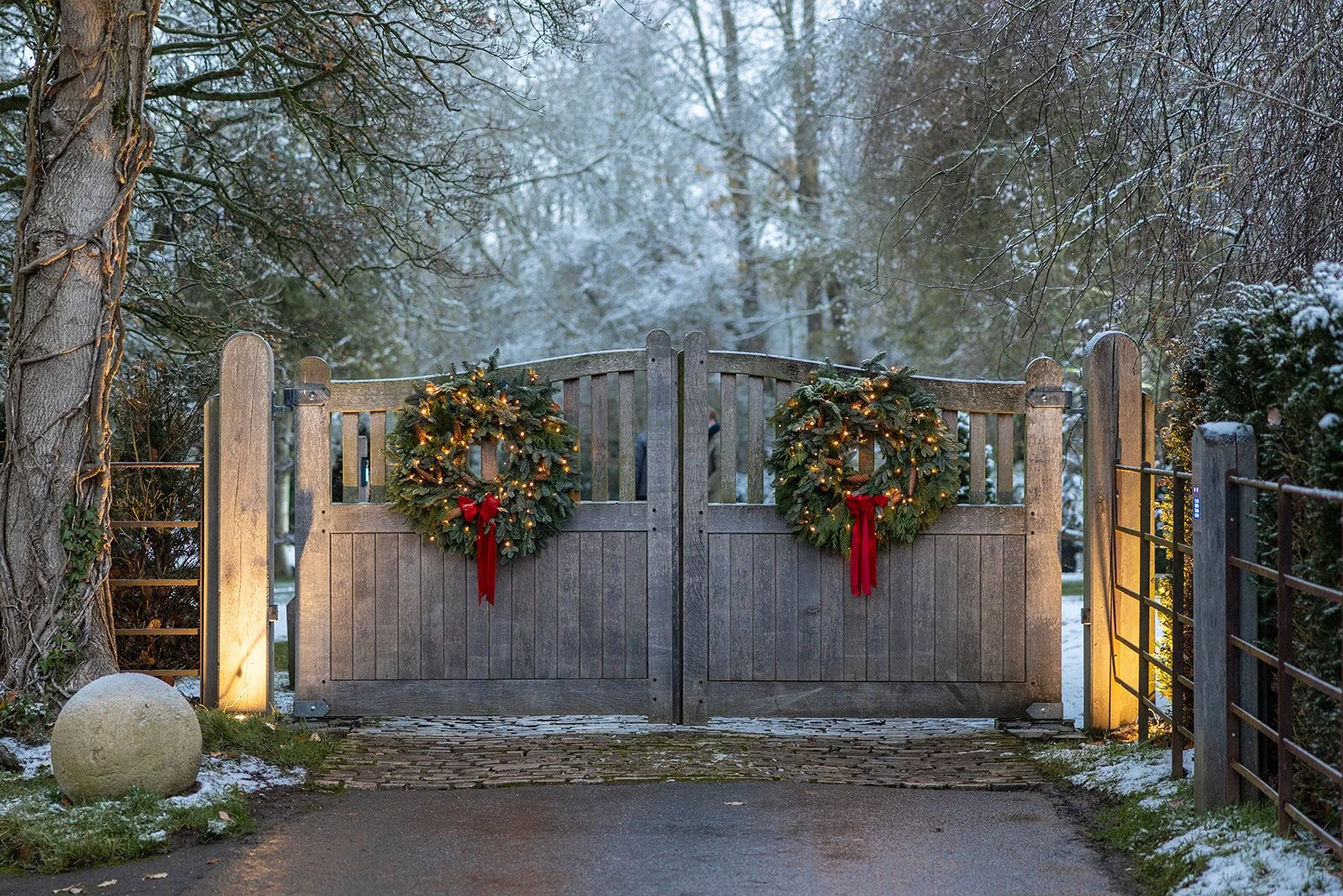 A wooden gate decorated with two Christmas wreaths with red bows and lights, set in a snowy outdoor scene with trees and shrubs, in the evening.