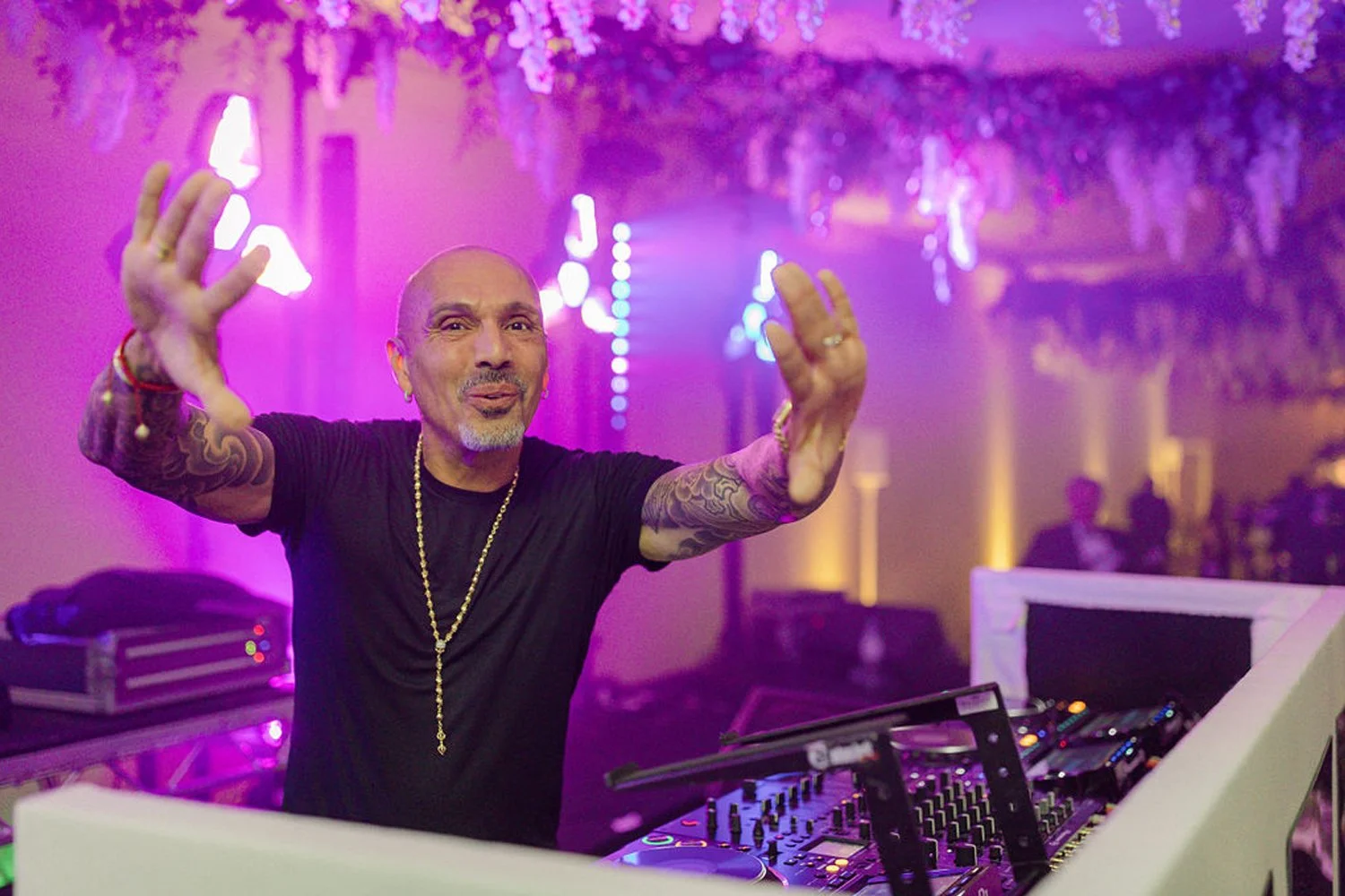 A DJ at a party or event with purple and pink lighting, gesturing with his hands towards the camera, wearing a black shirt, a gold chain, and tattooed arms.