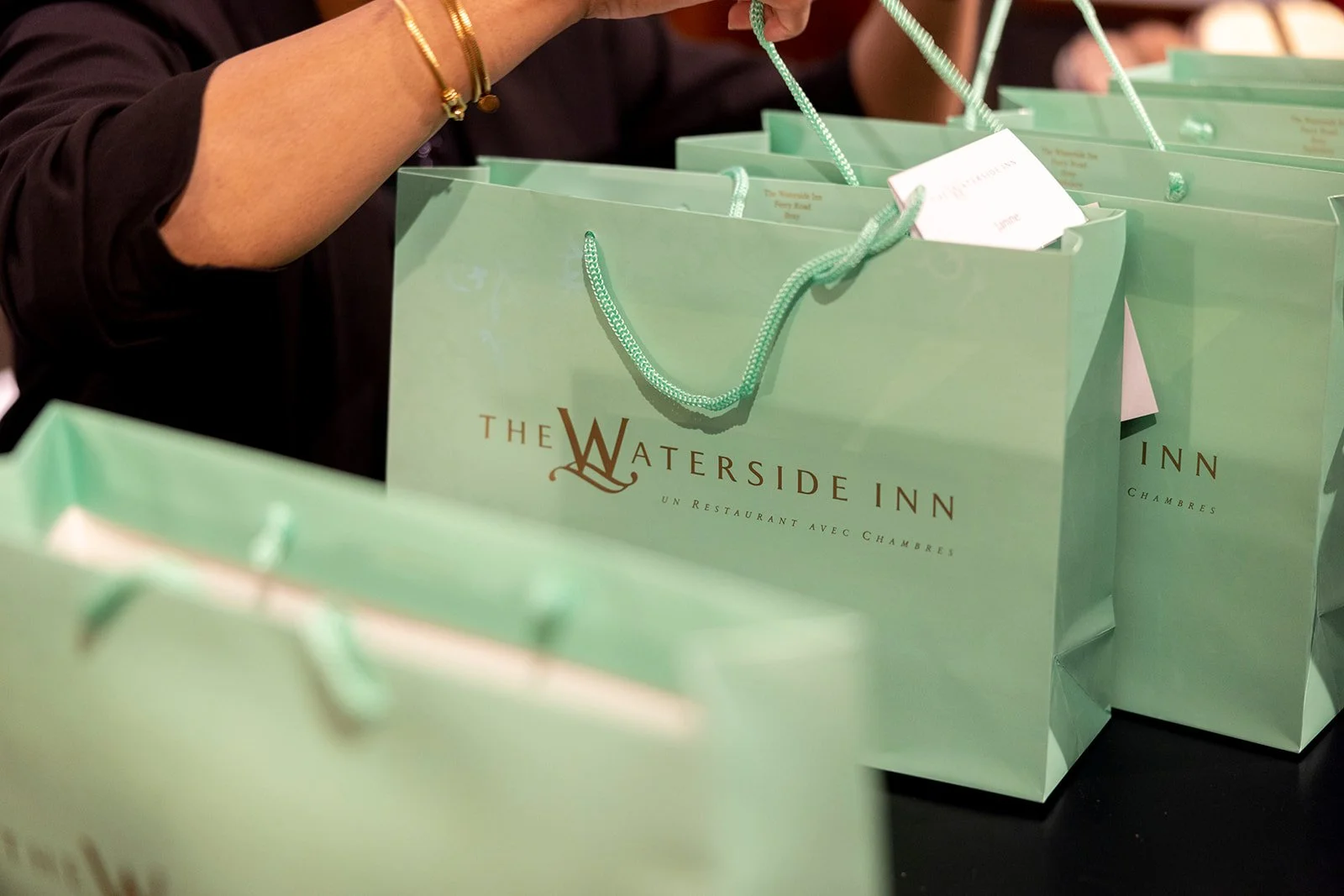 Light green paper shopping bags with gold and green handles, stacked on a table. The bags are from The Waterside Inn, an unrestaurant with rooms, printed in brown and gold lettering.