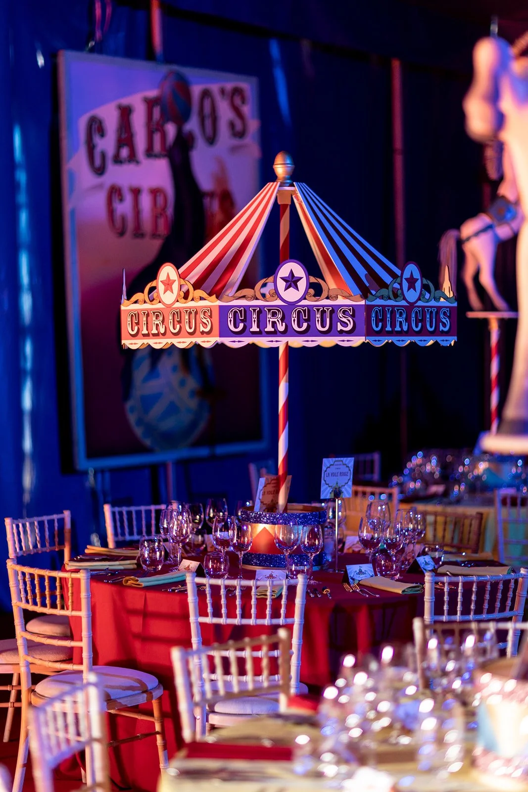 Colorful circus-themed table centerpiece with a striped canopy reading 'Circus' and a red table with glassware and chairs at a decorated event.