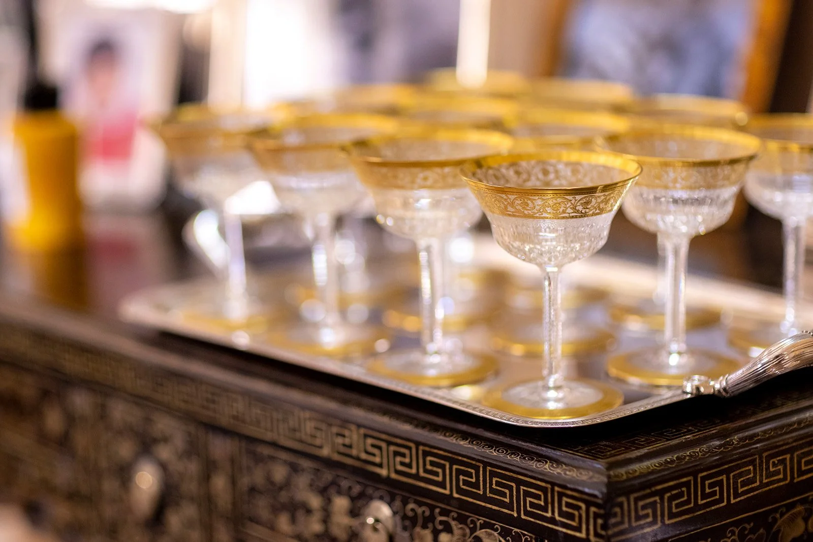 A tray of elegant, vintage glass coupe wine glasses with gold detailing around the rims, arranged on an ornate decorative surface.