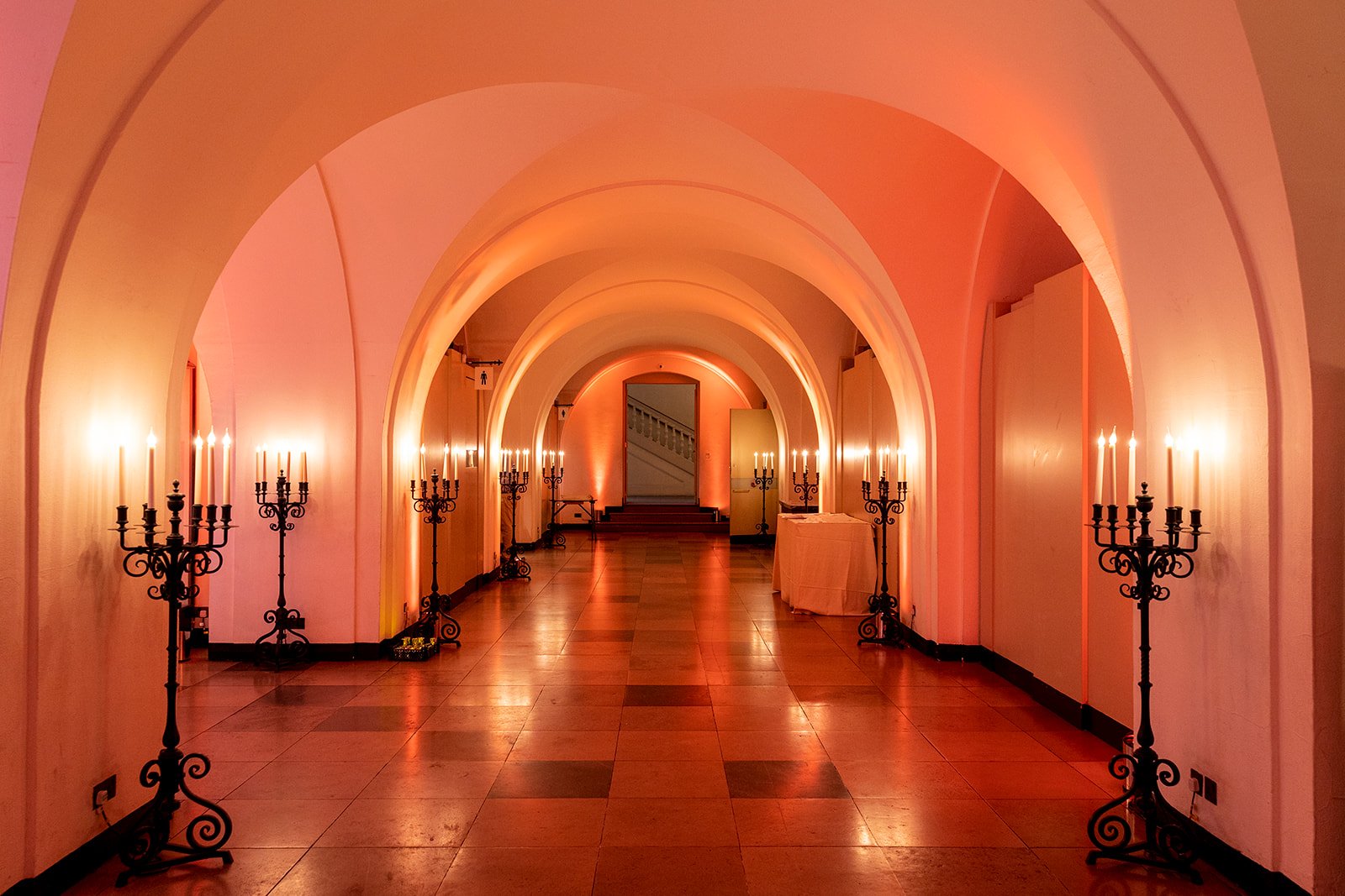 Elegant hallway with arched ceilings, warm pink lighting, and black candelabra-style candle holders along both sides.