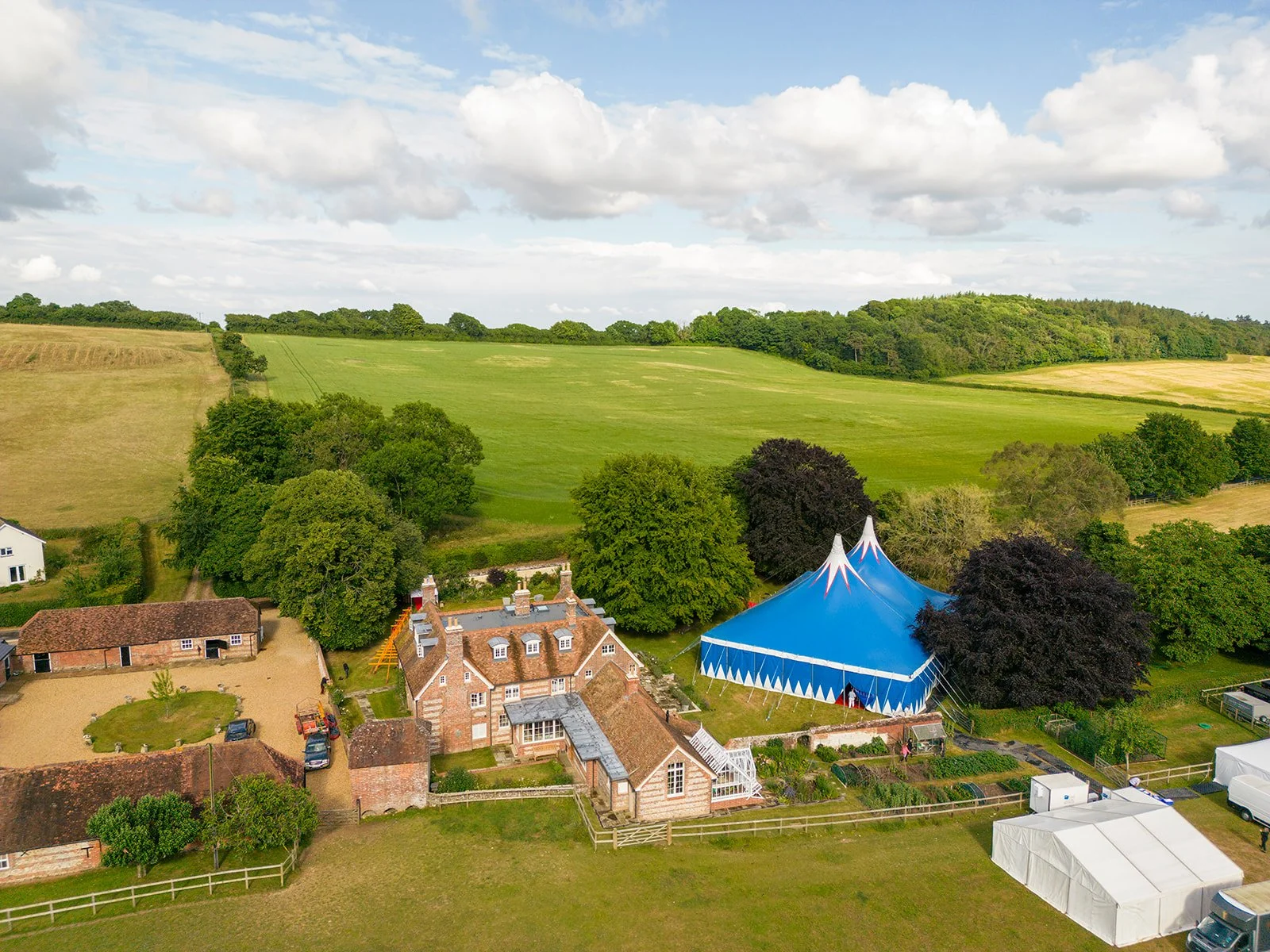 Aerial view of a rural landscape with green fields, trees, and a large blue circus tent near houses.