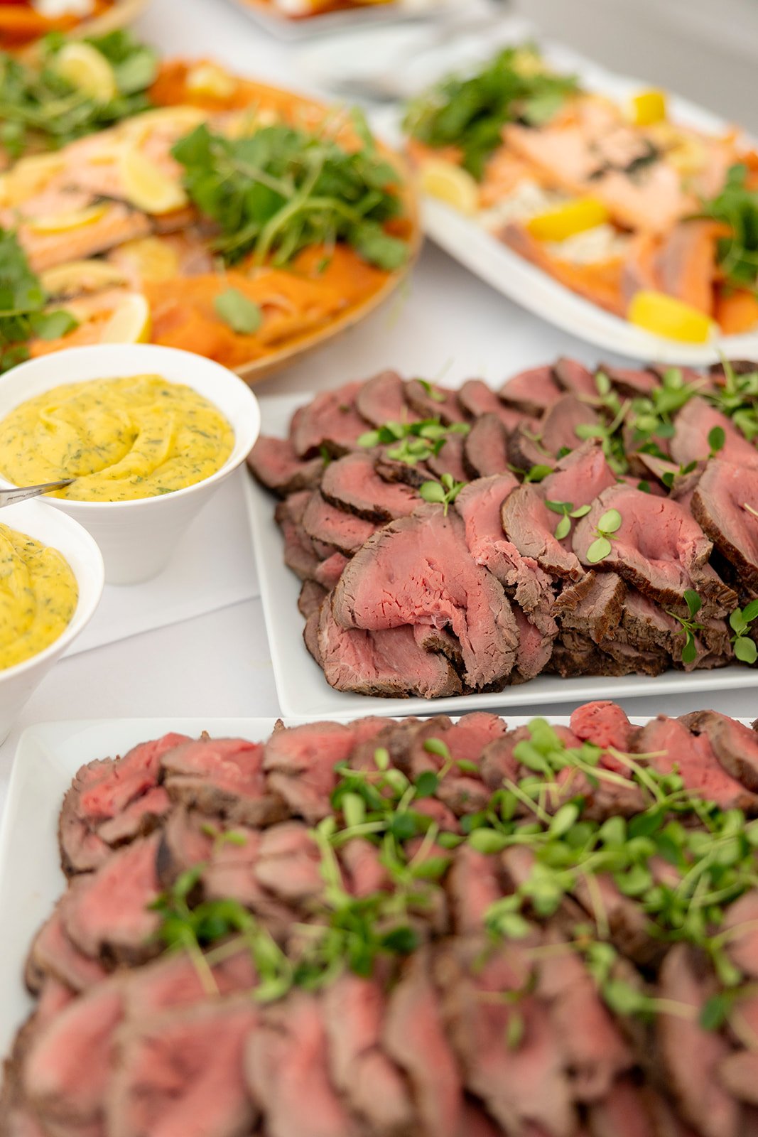 Sliced cooked steak garnished with microgreens on a white platter, with bowls of mustard sauce nearby, and other dishes like salad and salmon in the background.