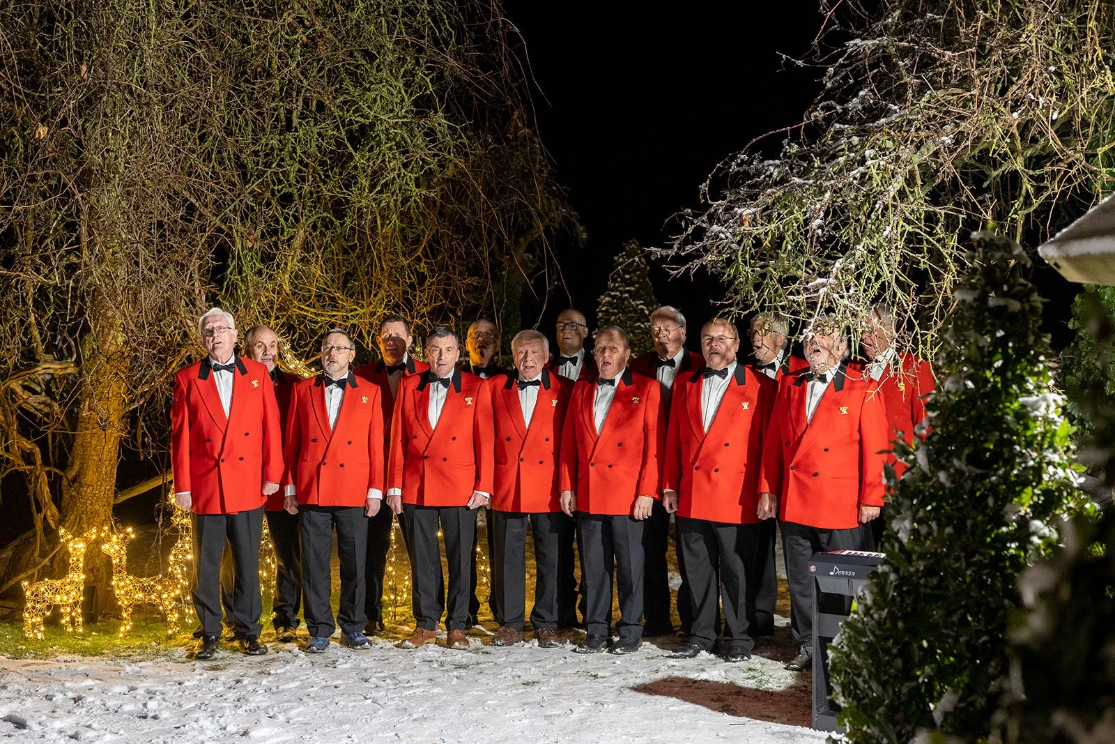 A choir of adult men dressed in red blazers, white shirts, black bow ties, and black pants, singing outdoors at night surrounded by leafless trees and Christmas lights.