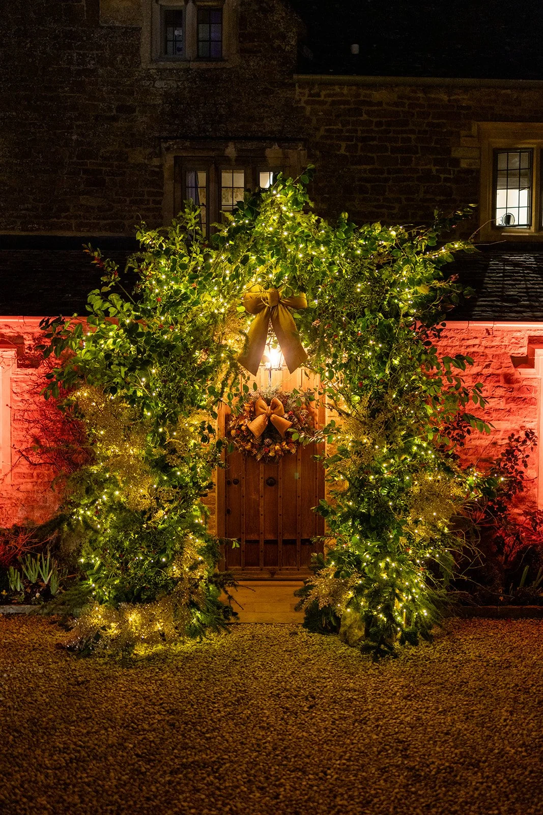 Decorated wooden door with a Christmas wreath, surrounded by greenery and string lights, and a large gold bow on top, with a brick house wall and windows in the background.