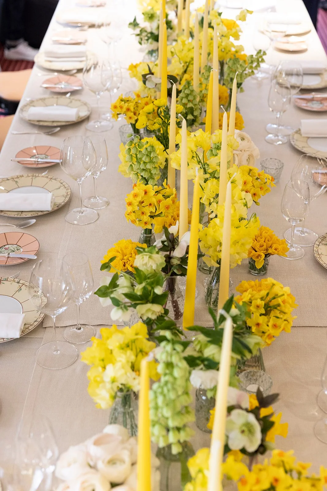 Elegant table setting with a beige tablecloth, yellow floral centerpiece, tall yellow candles, and fine china plates with cloth napkins, surrounded by glasses and silverware for a formal event.
