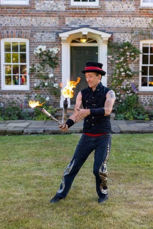 A man wearing a black sleeveless shirt, black cap, and jeans, performing a fire juggling act outdoors in front of a brick house with white windows and garden.