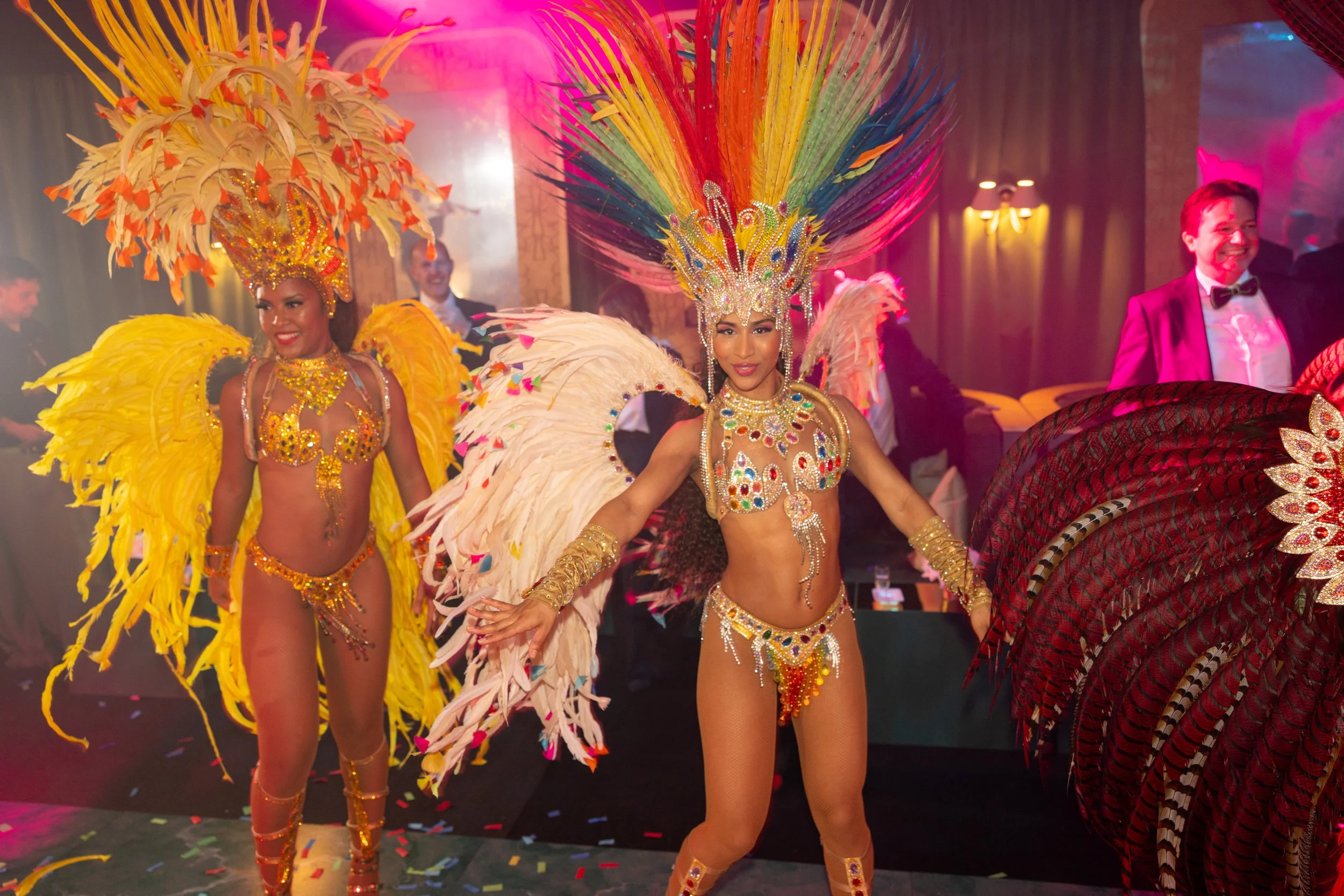 Dancers dressed in colorful carnival costumes with large feathered headdresses and jewel embellishments, dancing in a lively indoor setting.