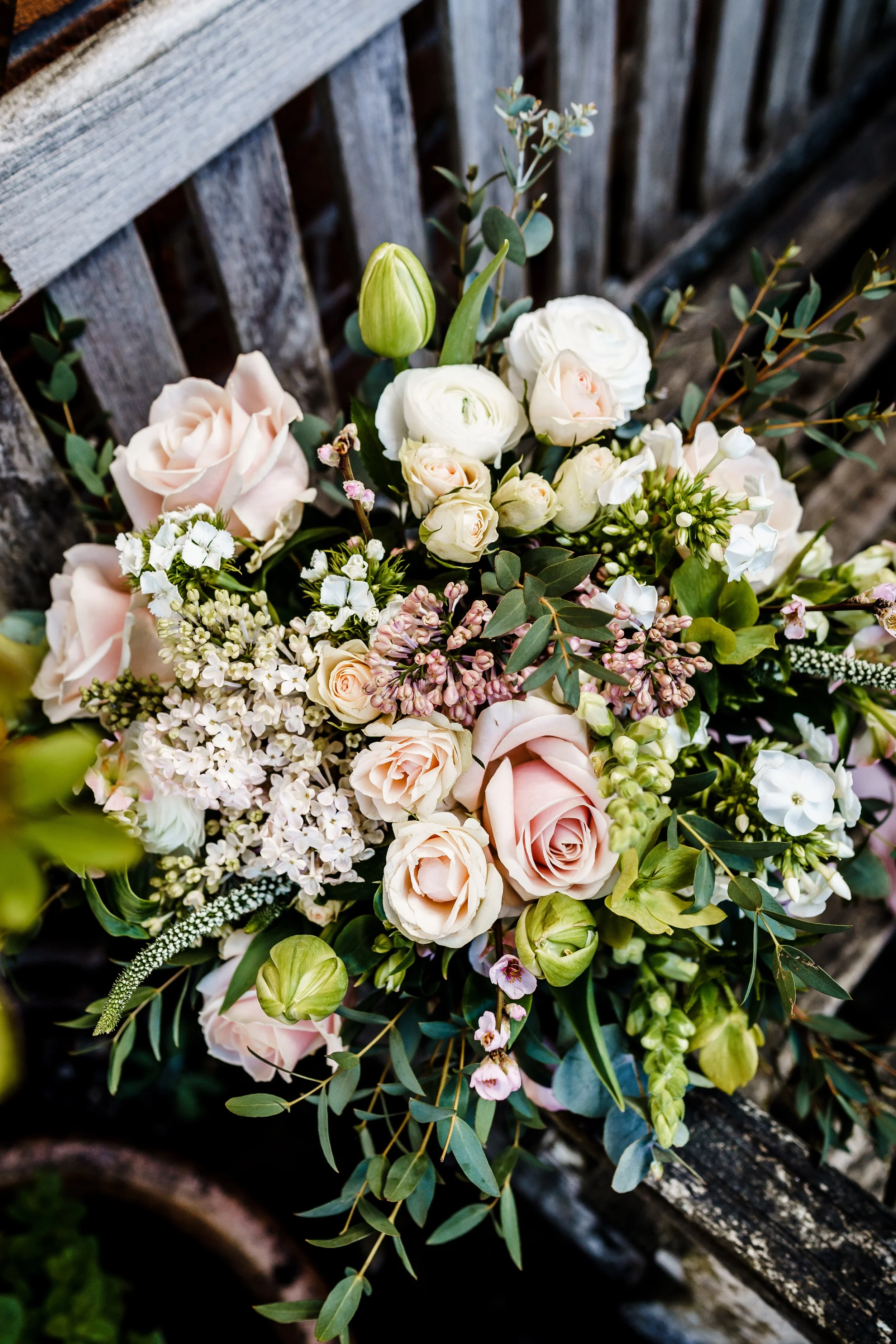 A colorful flower arrangement on a wooden surface with greenery and flowers including roses, small white flowers, pink buds, and eucalyptus leaves.