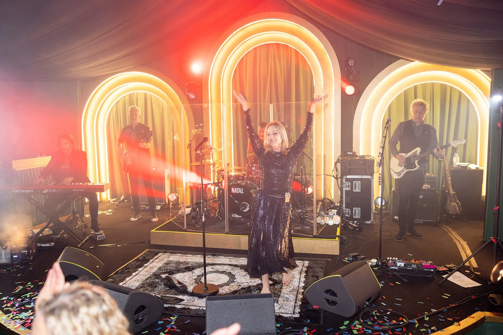 A woman in a black sequined dress performing on stage with a band, surrounded by decorative arches with neon lighting, music equipment, and colorful confetti on the floor.