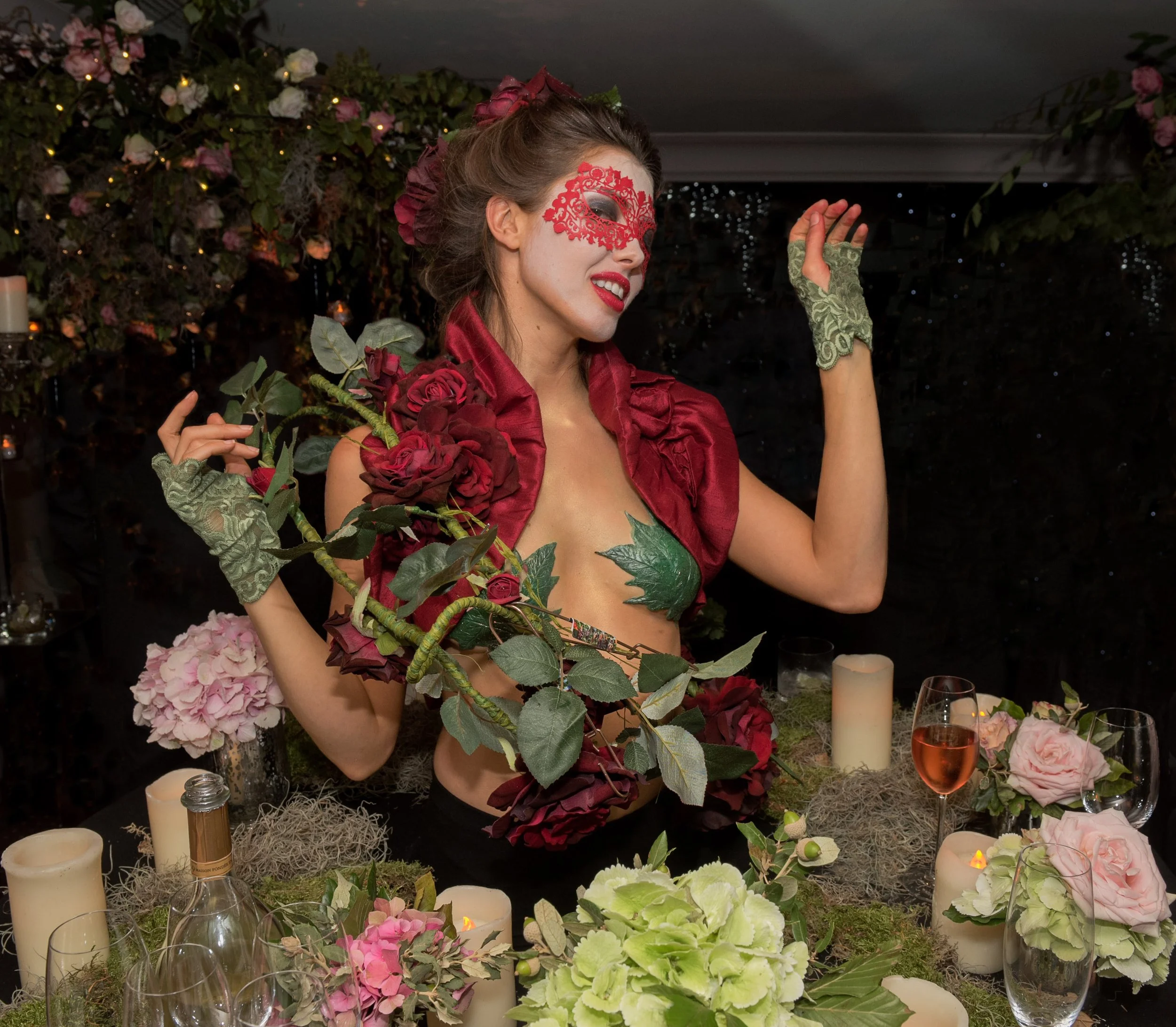 A woman dressed in a red outfit with floral details, wearing a red lace mask, at a floral-themed event or celebration, surrounded by candles, flowers, and glasses of rosé wine.