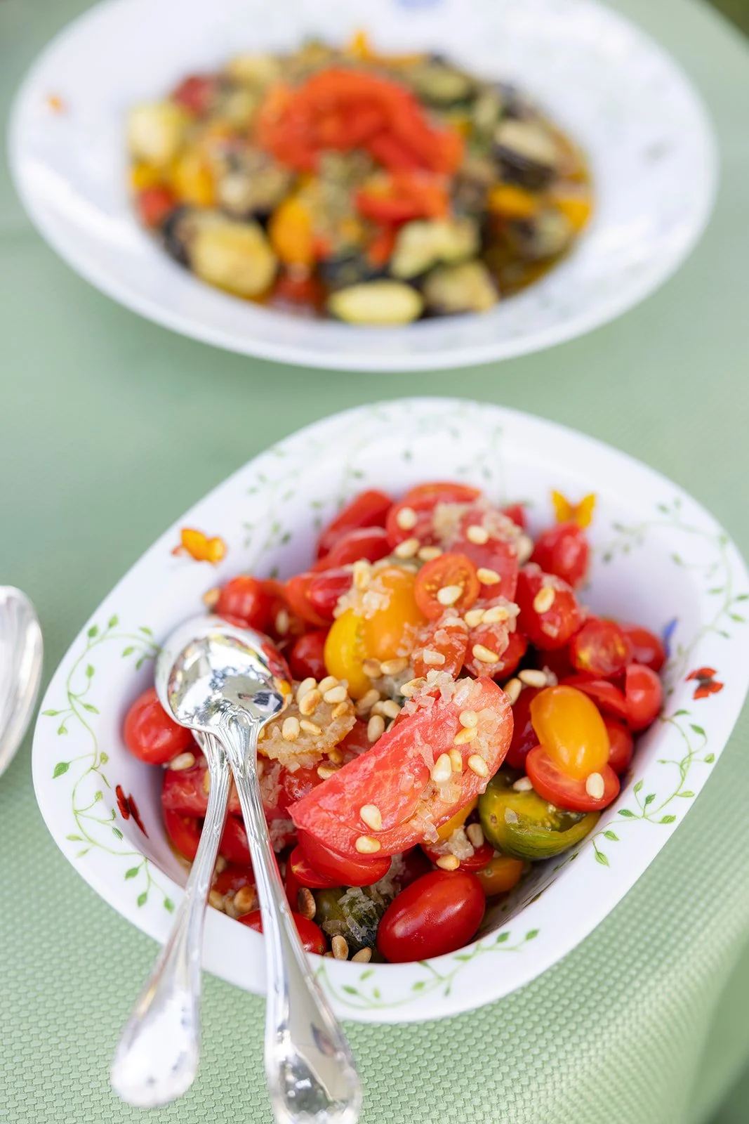 A white dish with a green vine pattern filled with a fresh tomato salad, topped with pine nuts and a drizzle of oil. In the background, another white bowl holds a cooked vegetable dish with red and yellow peppers and other vegetables.