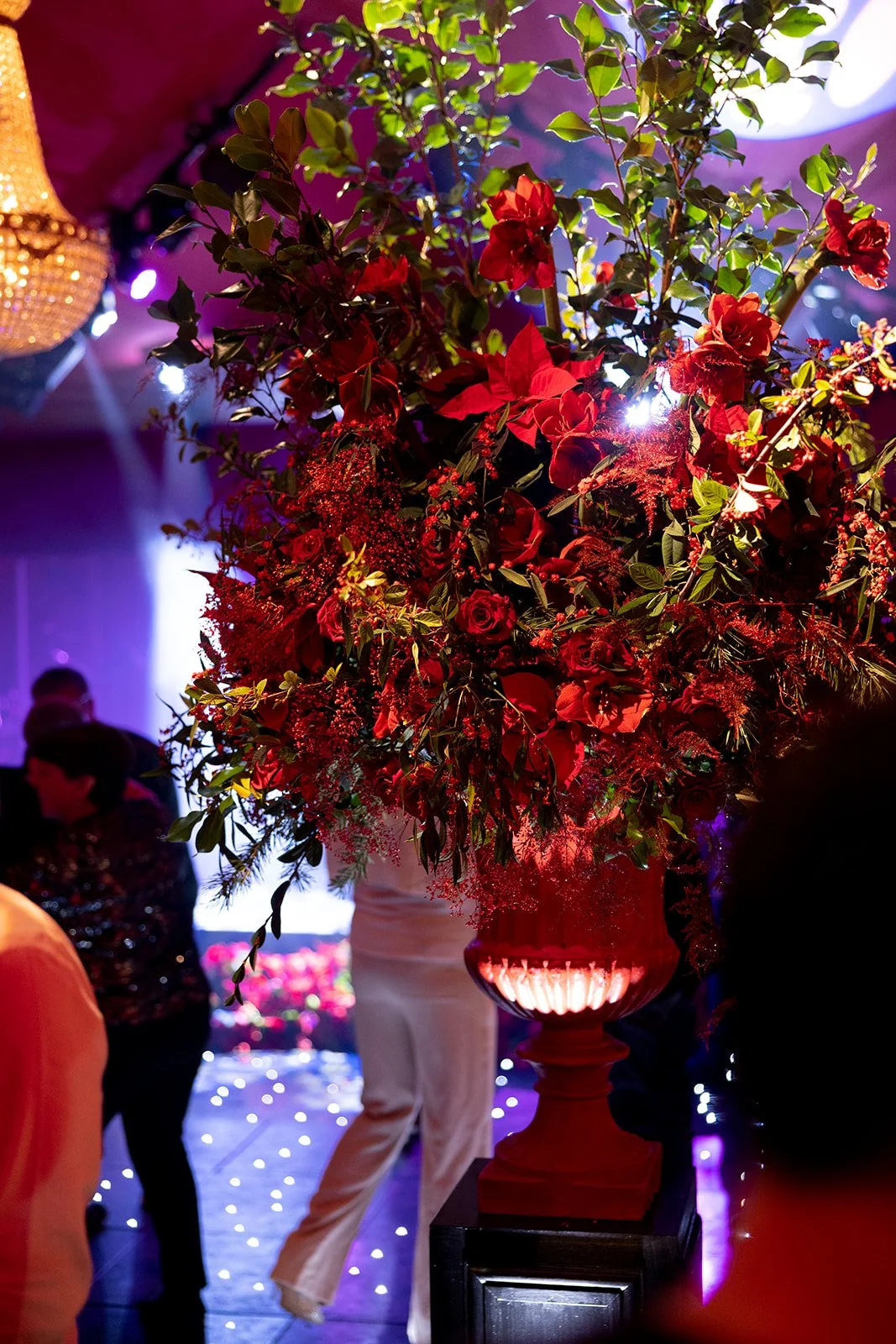 A large floral arrangement of red flowers and green leaves in a tall red vase with lighting underneath, set in a decorated event space with purple and blue lighting and blurred guests in the background.