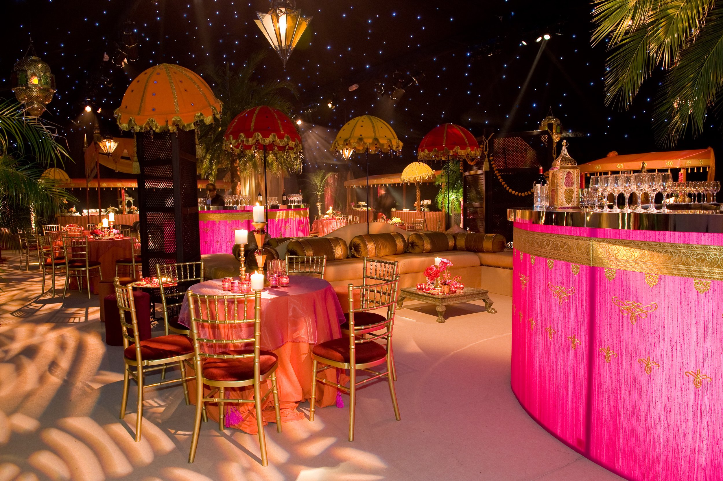 Decorative event space with pink and gold themed tables, ornate umbrellas, candles, lush greenery, and starry ceiling lighting, creating an elegant ambiance.