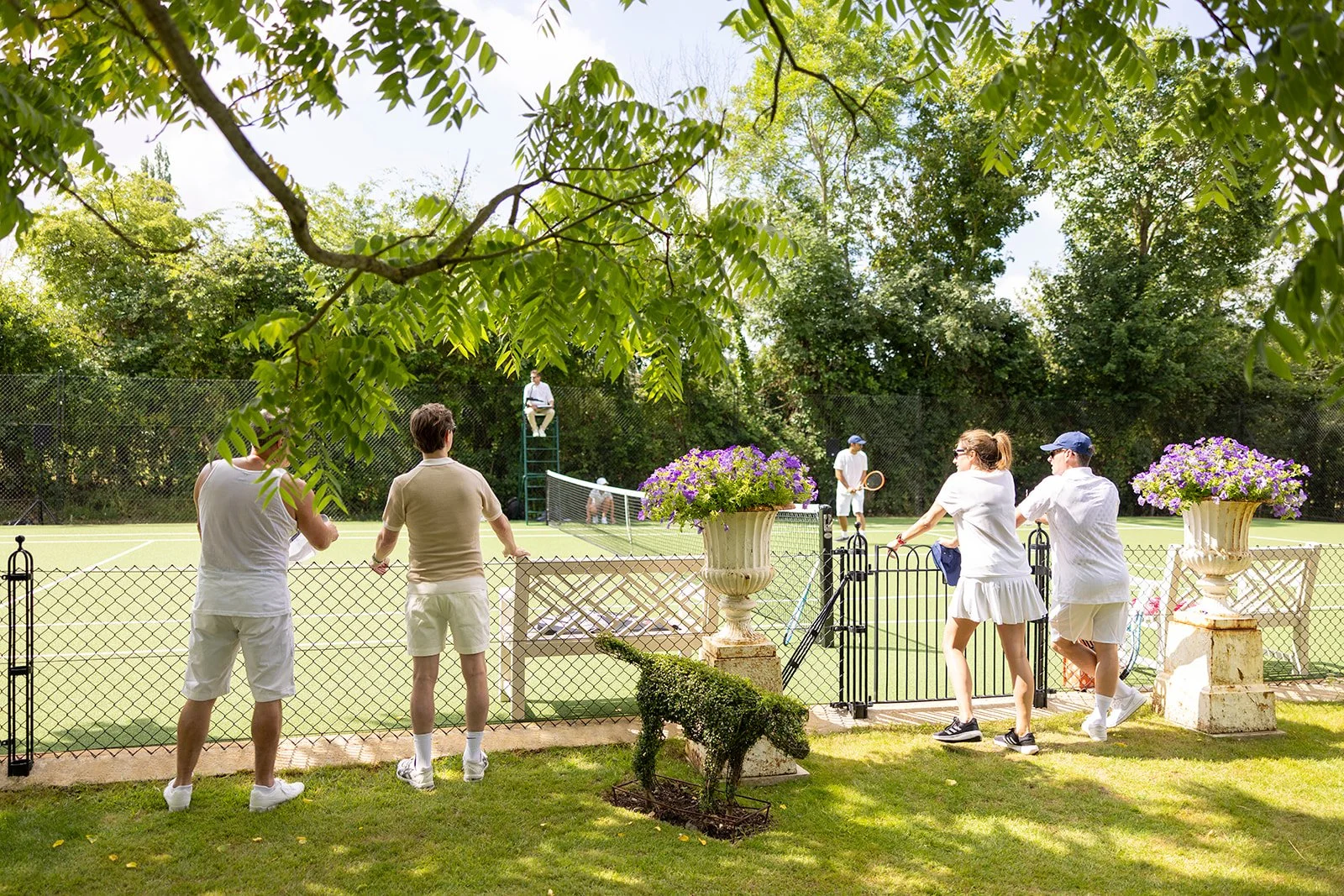 People playing tennis on a court surrounded by a fence and greenery, with a judge sitting on a high chair in the background and large flower pots with purple flowers near the gate.