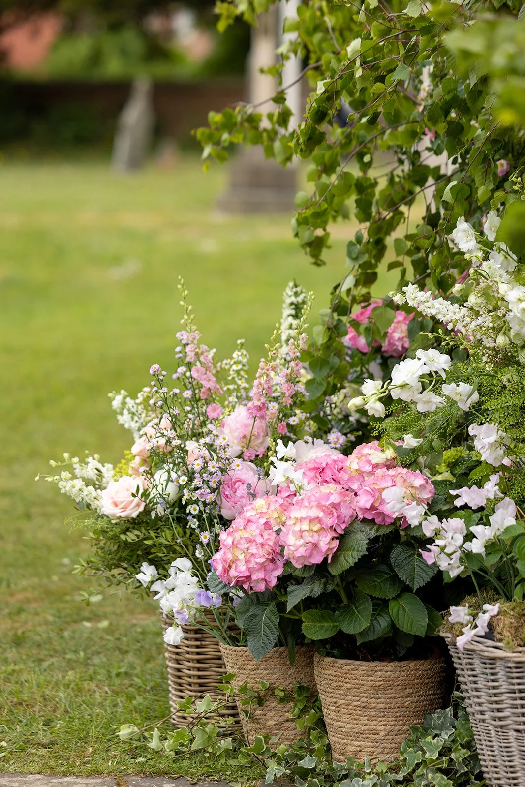 A garden scene with pink, white, and purple flowers in wicker baskets, with greenery and a blurred background.