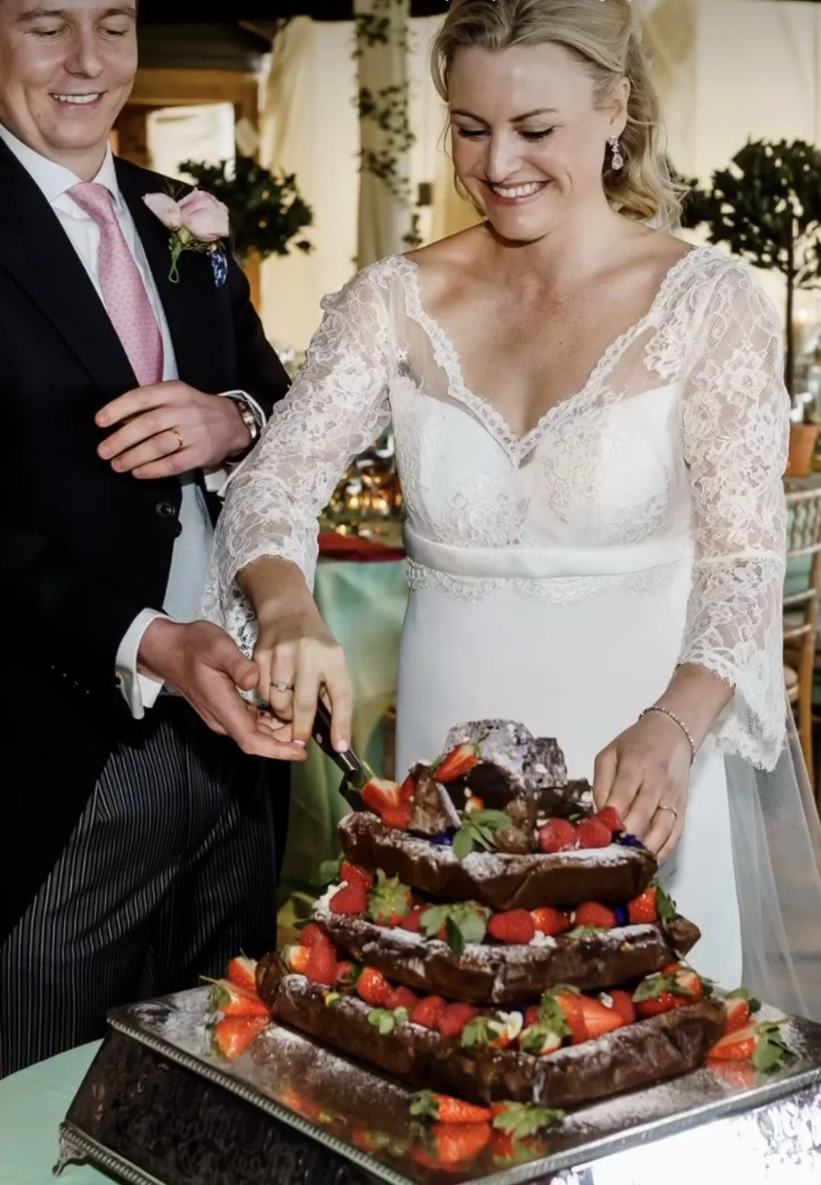 A bride in a lace wedding dress is cutting a wedding cake with strawberries and berries on top. A groom in a black suit and pink tie stands nearby, smiling.