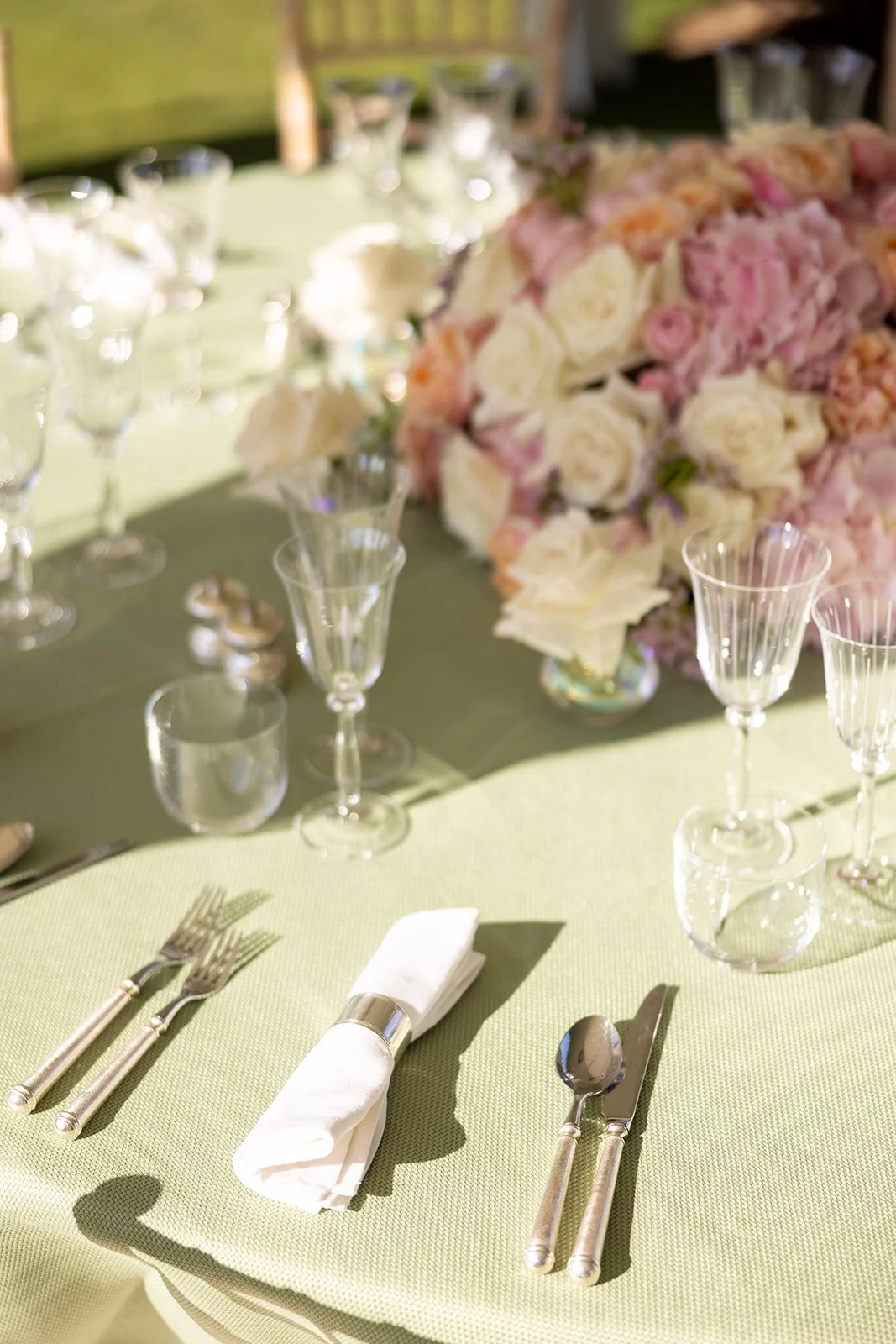 A close-up of a beautifully set outdoor dining table with glassware, silverware, a white napkin, and a floral centerpiece with pink and white flowers on a green tablecloth.