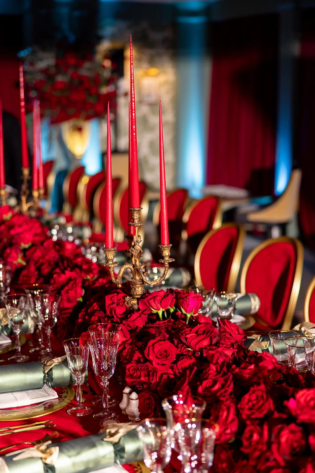 Elegant banquet table decorated with red roses, a gold candelabrum with red candles, and gold-rimmed glassware, set for a formal event with red and gold chairs in the background.