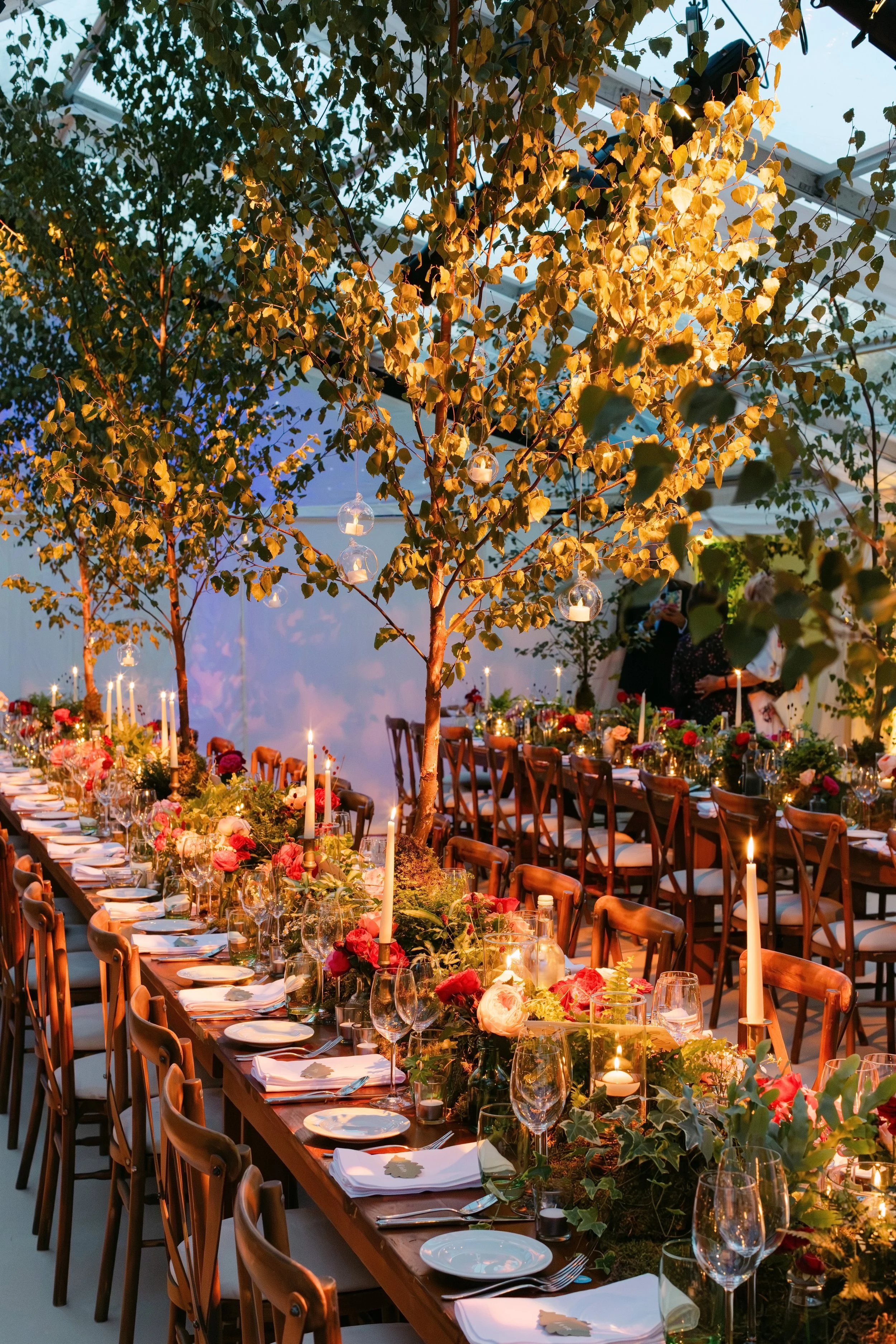 A long banquet table decorated with flowers, candles, and glassware, set up under hanging trees with hanging glass candle holders and string lights, in a brightly lit indoor space at dusk.
