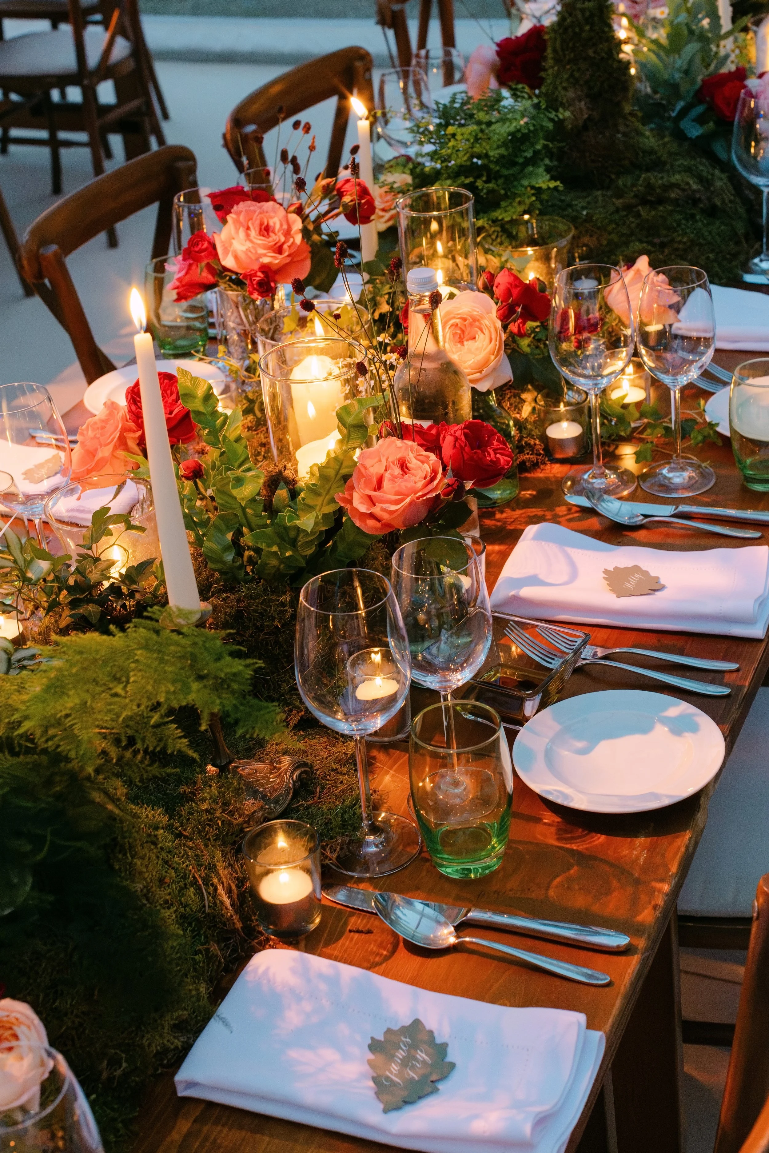 A beautifully decorated dinner table with pink roses, red flowers, candles, and greenery centerpiece, set with white napkins, wine glasses, and silverware for a formal event at dusk or evening.