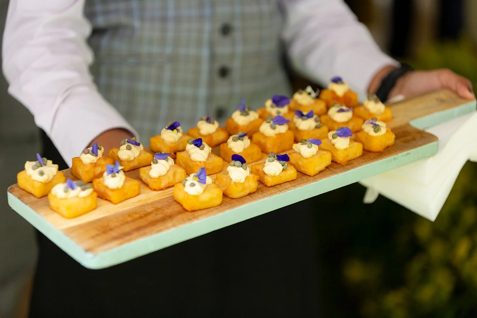 A person holding a wooden serving tray with small, square, fried appetizers topped with cream, purple edible flowers, and small green herbs.