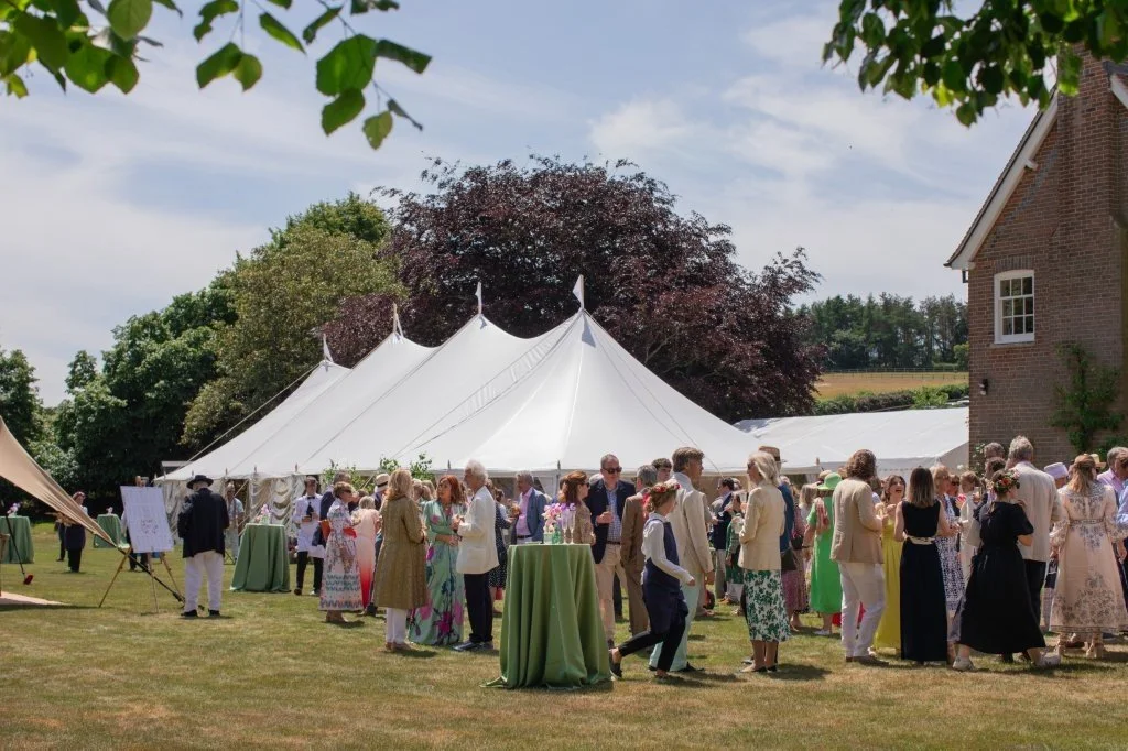 People attending an outdoor event with a large white tent, under trees and on a grassy area, with some wearing fancy or vintage clothing during daytime.