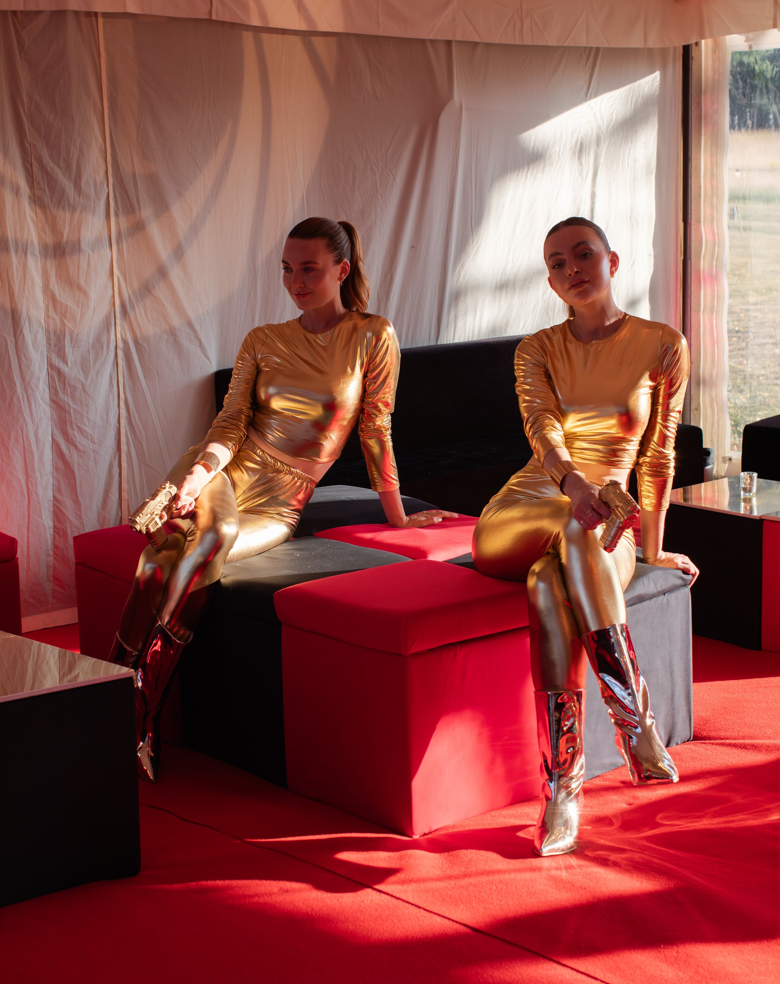 Two women dressed in shiny gold outfits with high boots sitting on a black and red sofa in a brightly lit room with draped curtains and sheer walls, holding guns.